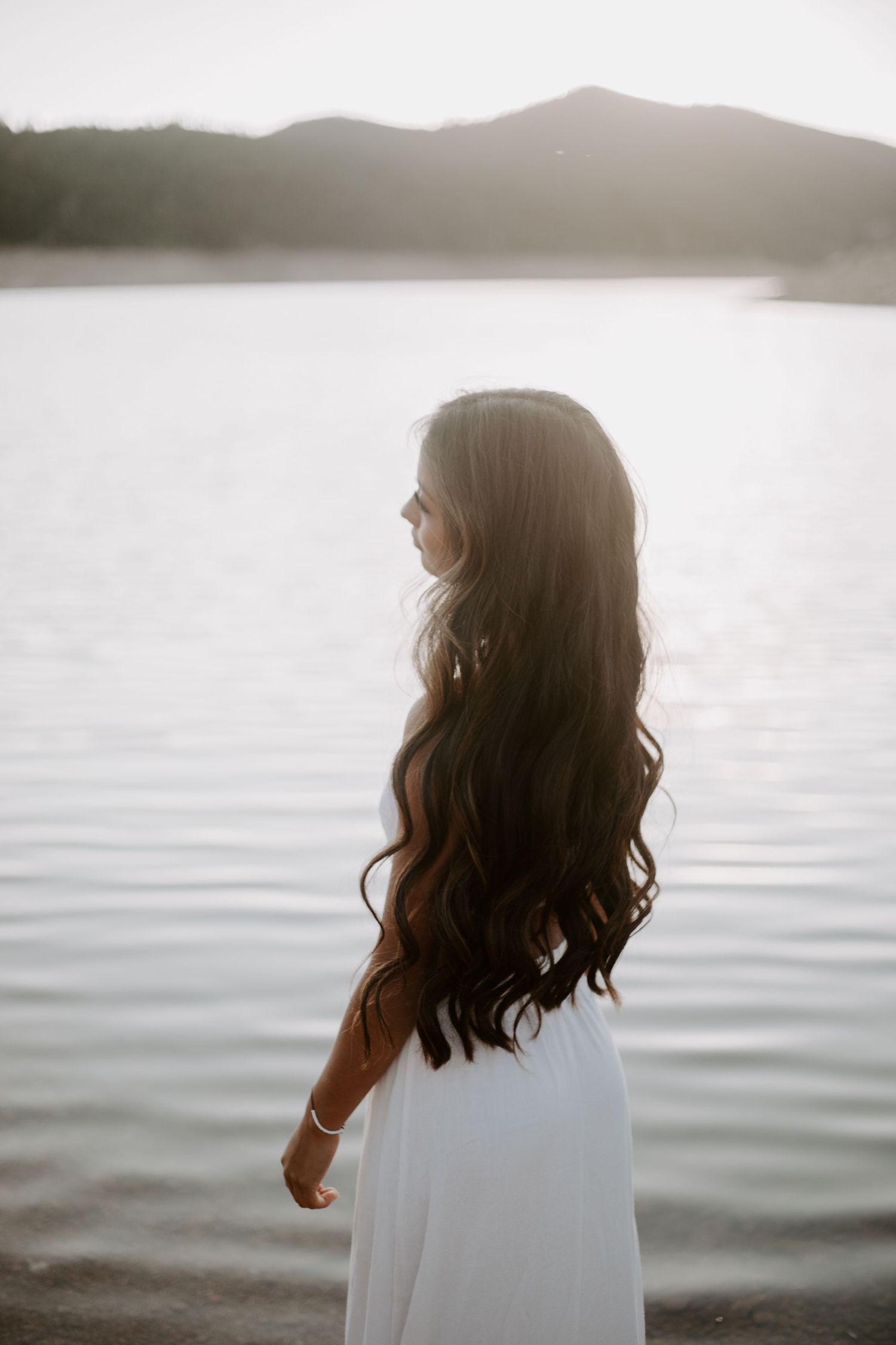 Woman with flowing dark hair in white dress standing by serene lake at sunset.