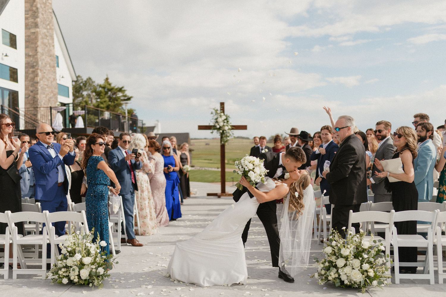 Bride and groom share a romantic dip kiss at outdoor wedding ceremony with white floral arrangements and cross altar.