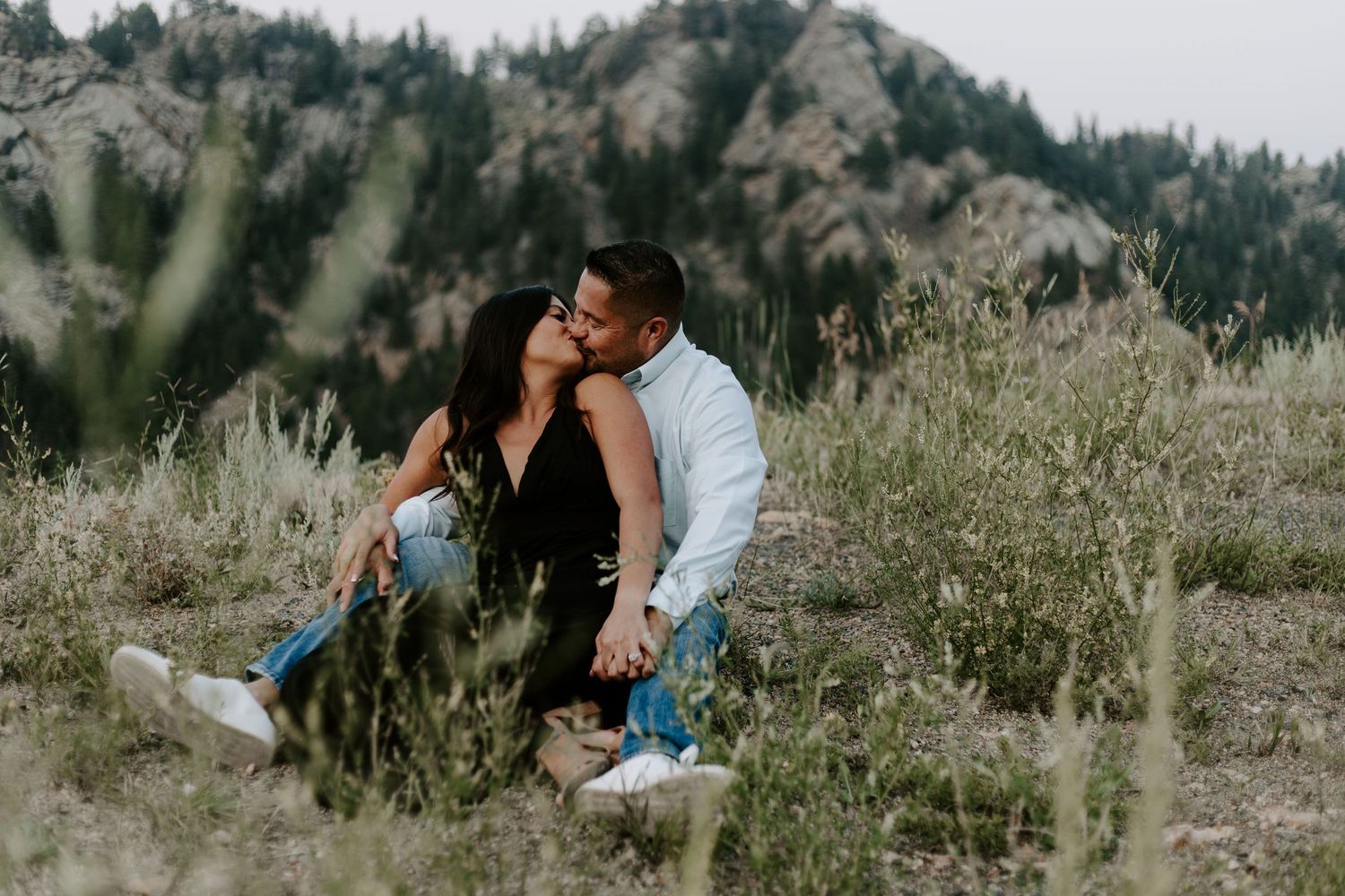 A couple shares an intimate moment while sitting in tall grass on a mountainside.