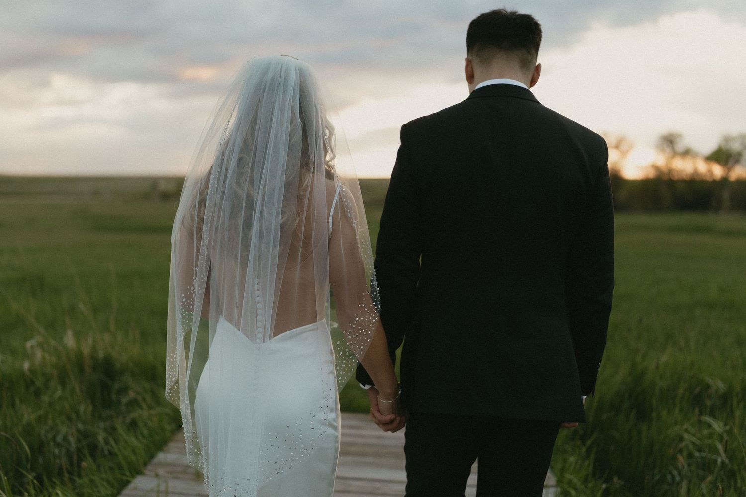 Bride and groom holding hands on a wooden path surrounded by green fields at sunset.