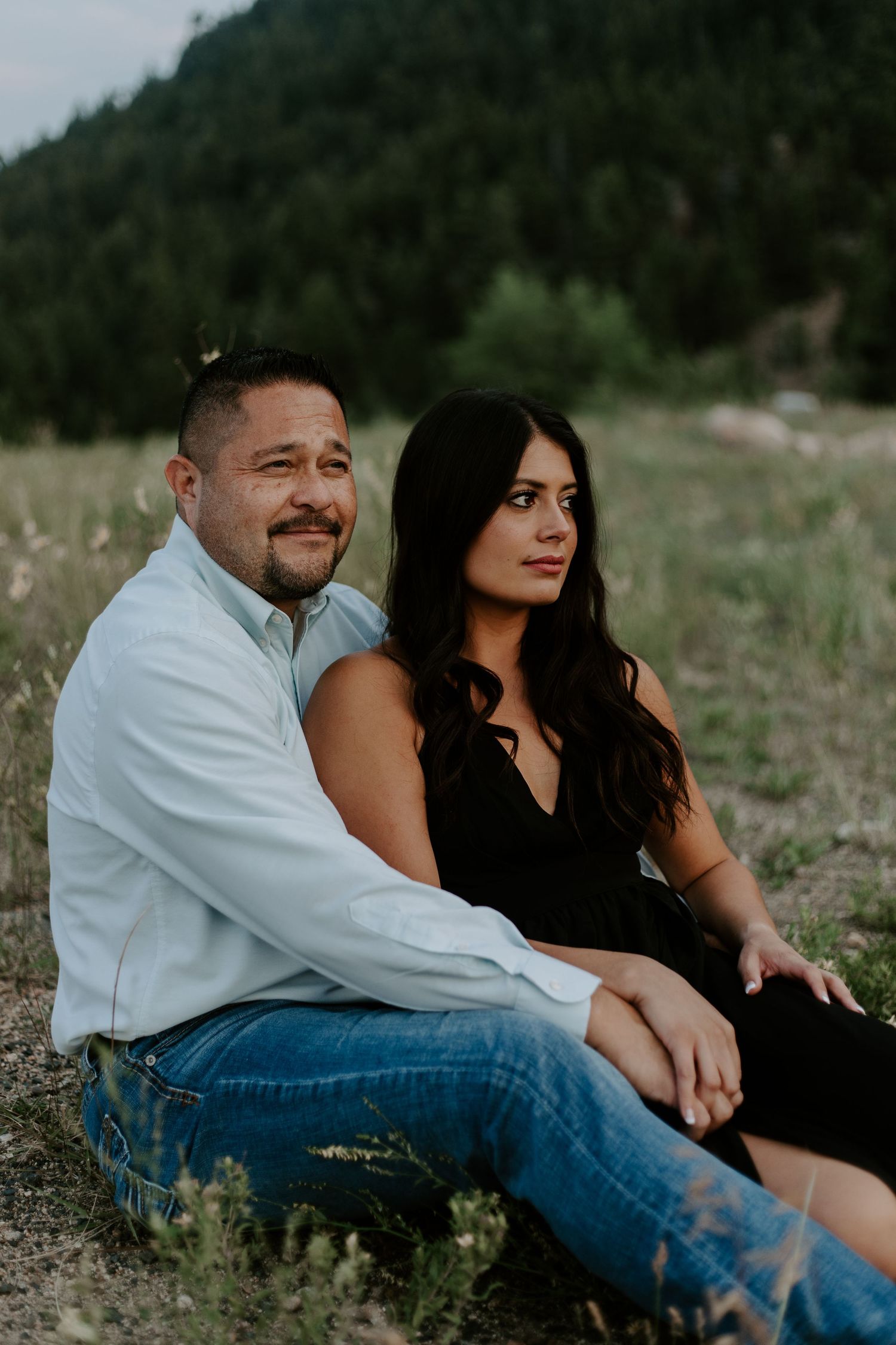 A couple sits together on a grassy hillside with mountains in the background during an engagement photoshoot.