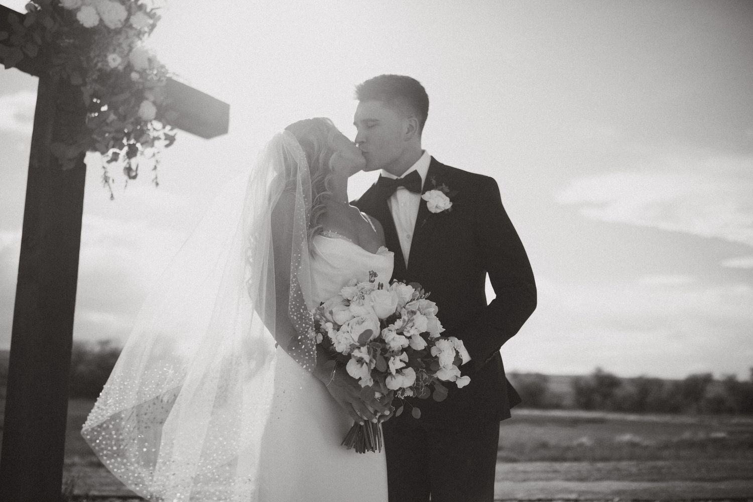 Black and white photo of a bride and groom sharing a kiss at their outdoor wedding ceremony.