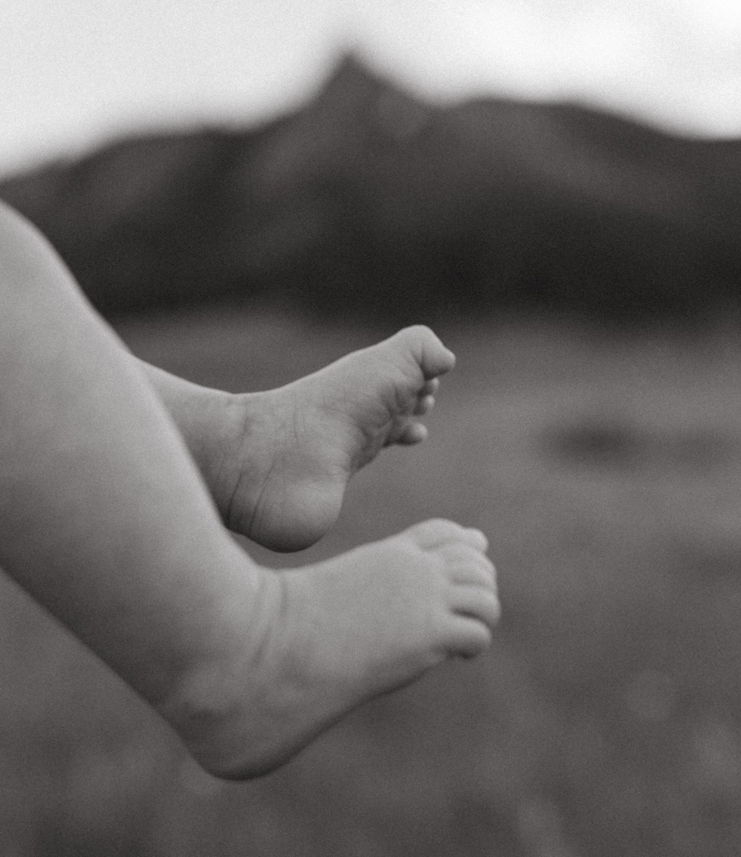 Black and white close-up of baby feet against mountain landscape background.