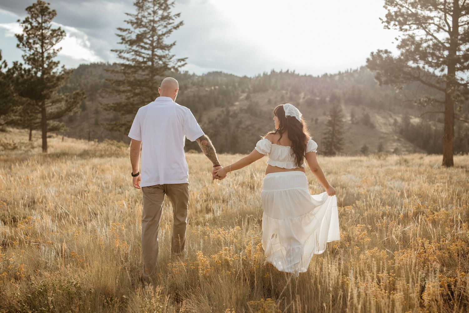 A couple walks hand in hand through a golden field at sunset with mountains and pine trees in the background.