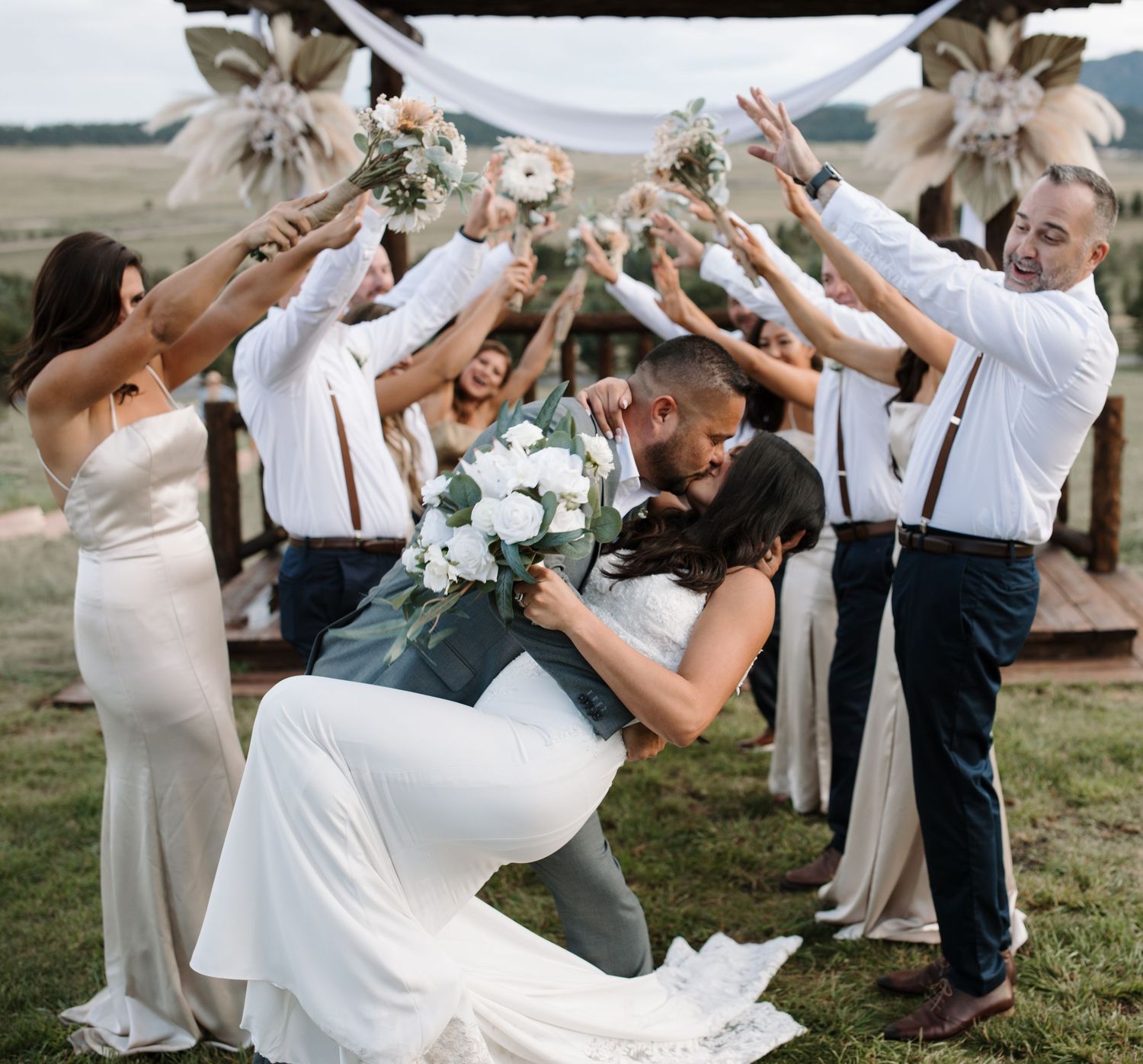 Wedding party celebrates outdoors with bridal couple sharing romantic dip kiss while guests cheer with raised hands.