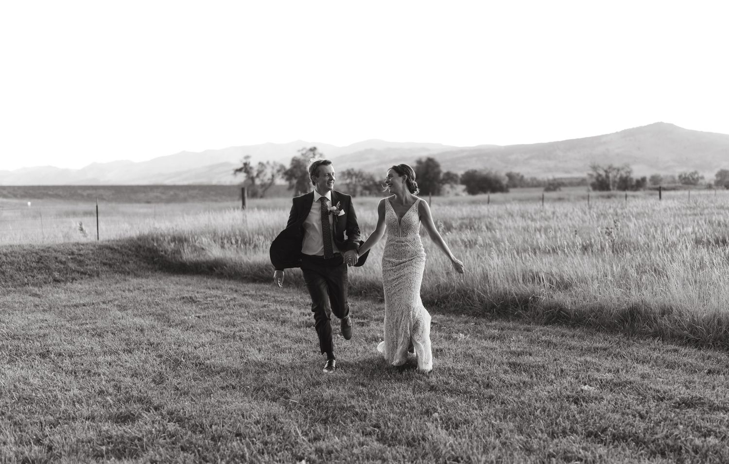 A black and white photo of newlyweds running joyfully through an open field.