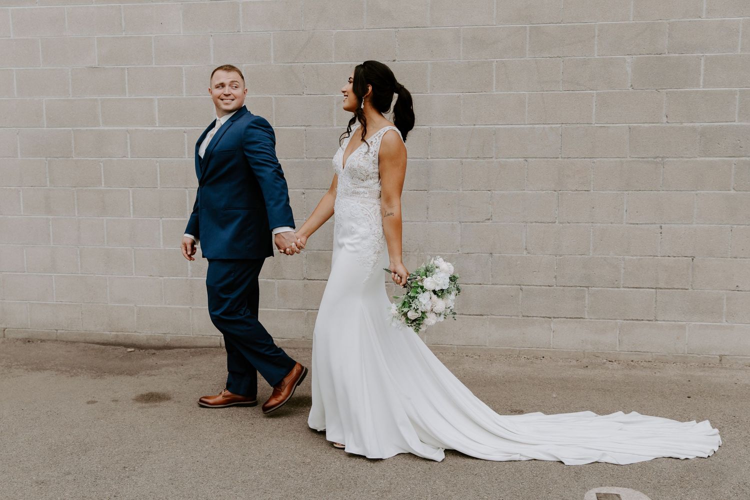 Wedding couple walking hand in hand along a concrete wall in formal attire.