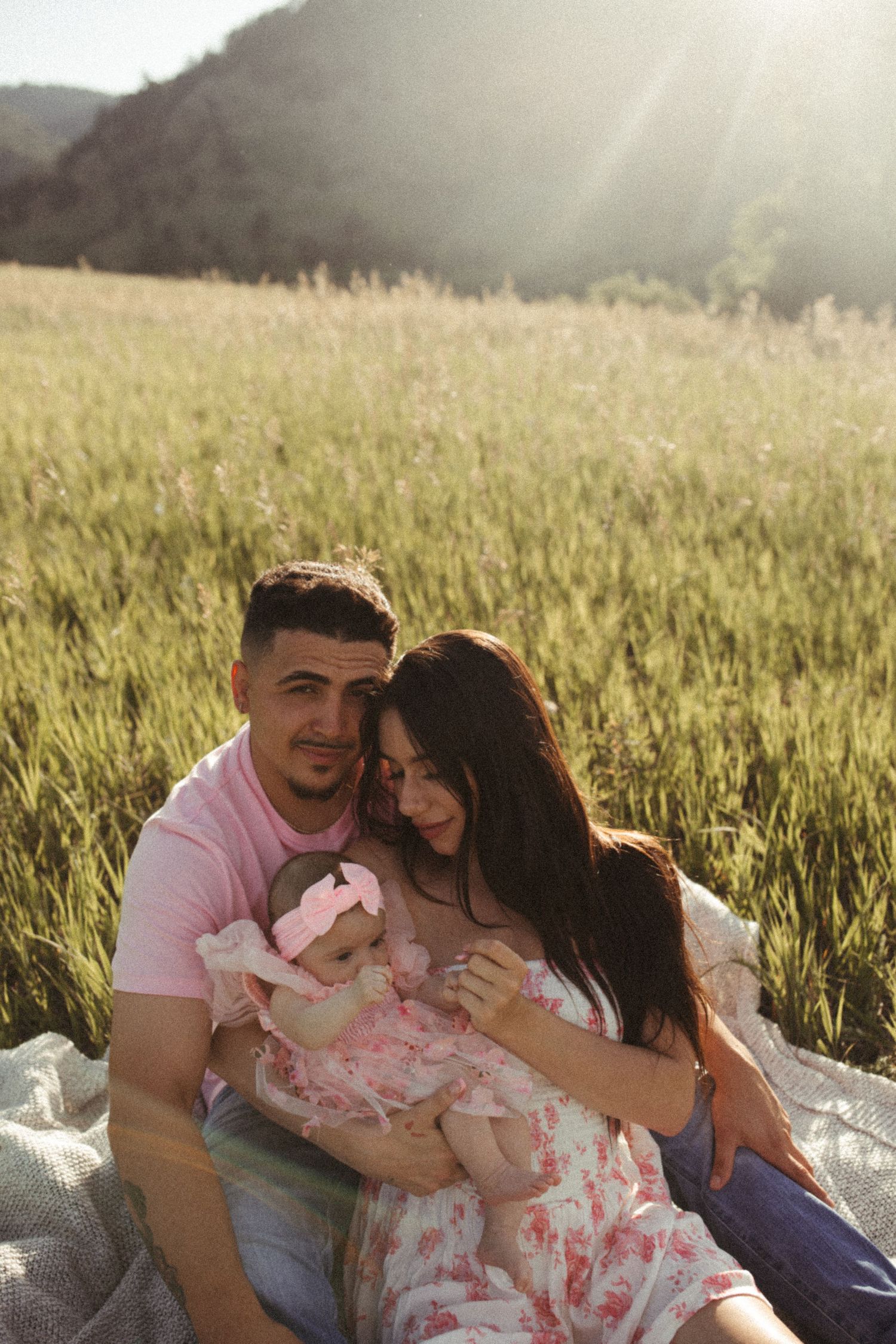 Family enjoying a tender moment together on a blanket in a sunlit meadow with mountains in background.
