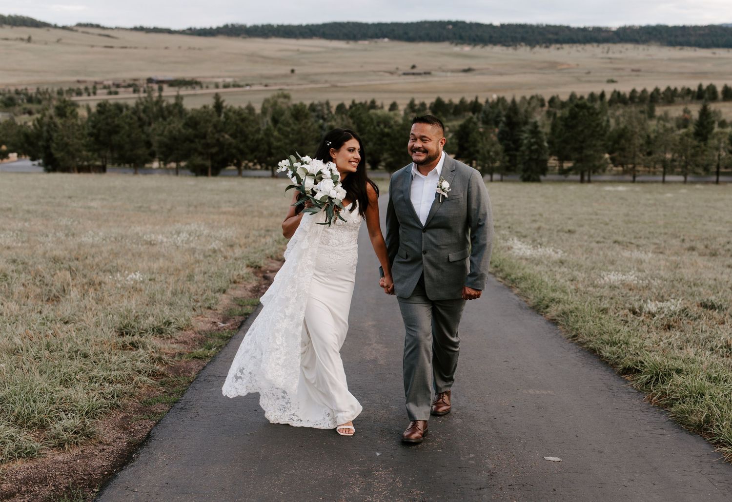 Newlyweds walking together on paved path through mountain meadow carrying white floral bouquet.