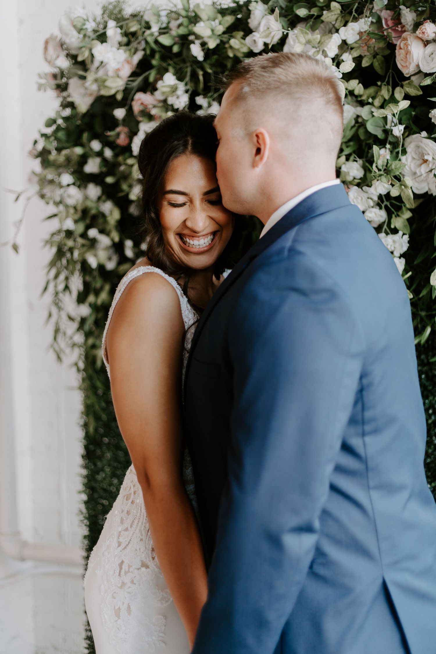 Intimate wedding moment captured against a floral arch backdrop with joyful laughter.