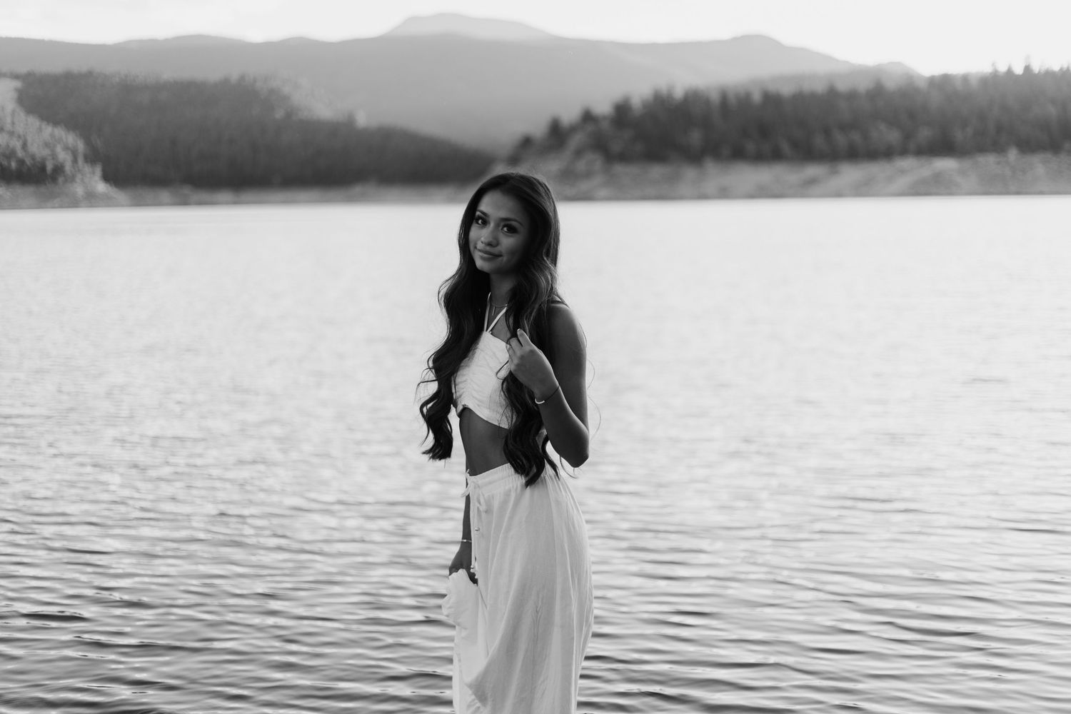 Black and white portrait by lake wearing flowing white dress with mountains in background.