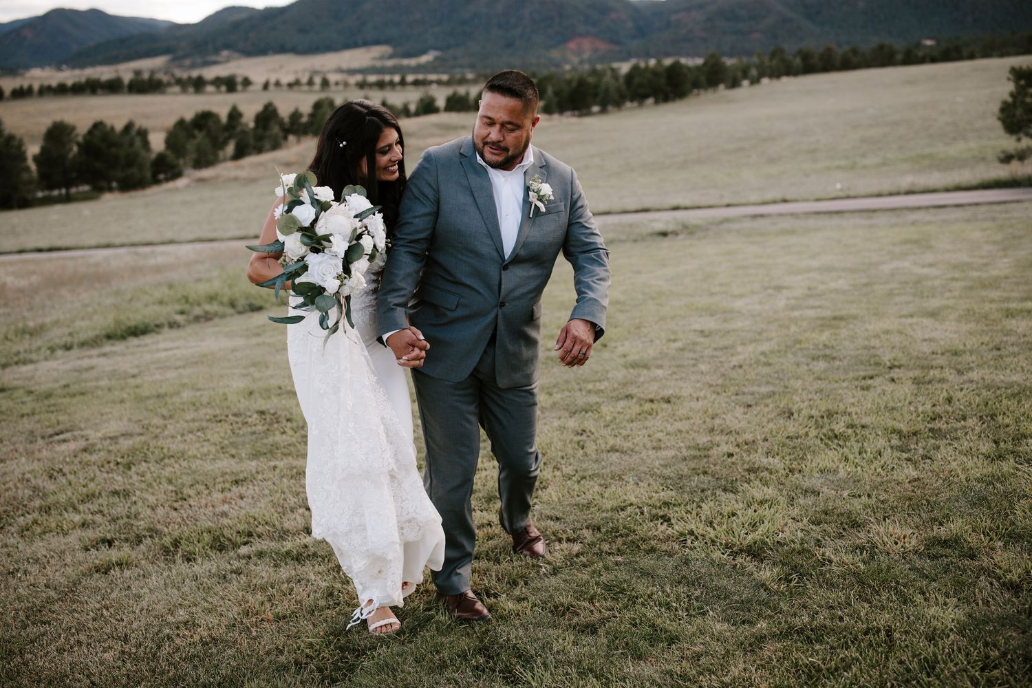 A romantic wedding couple walks together across a mountain meadow with the bride wearing a flowing white dress.