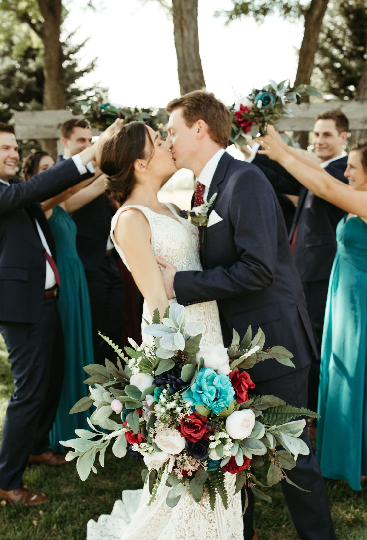 Wedding party celebrates with bouquets featuring red roses and greenery under tree canopy.