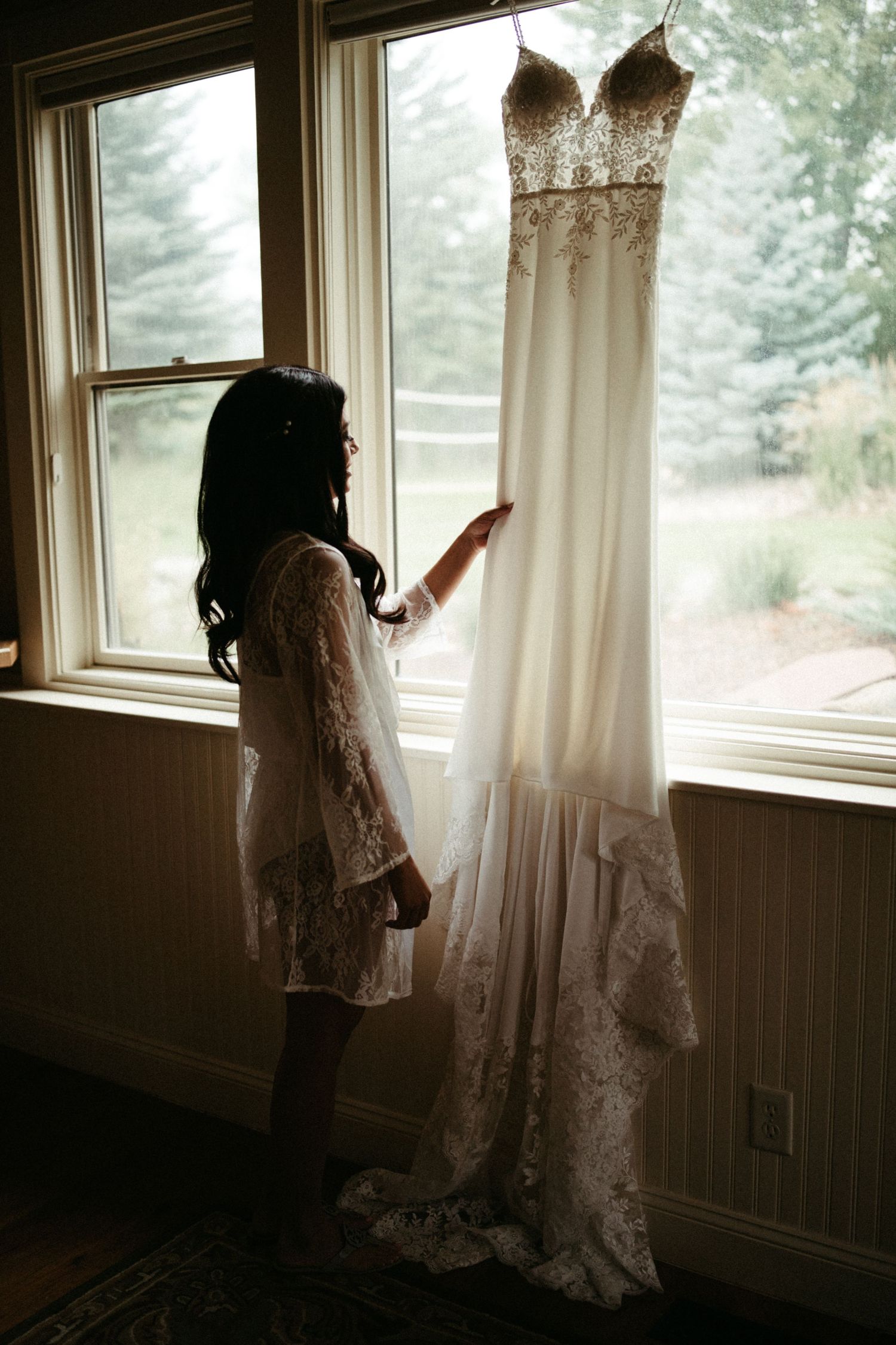 Silhouette of woman admiring elegant white lace wedding dress hanging by window.