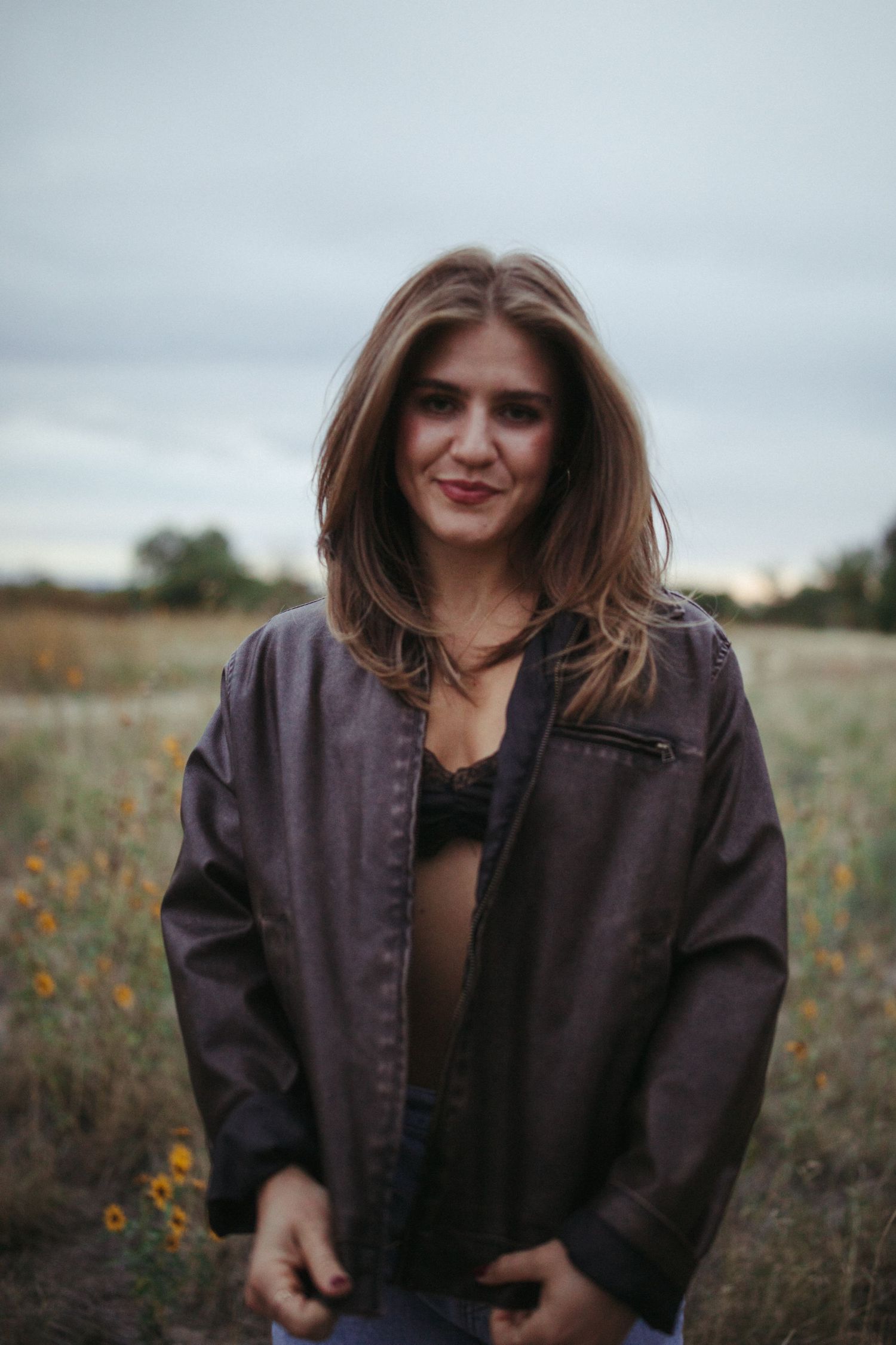 A person wearing a brown jacket poses in a field of wildflowers during an overcast day.