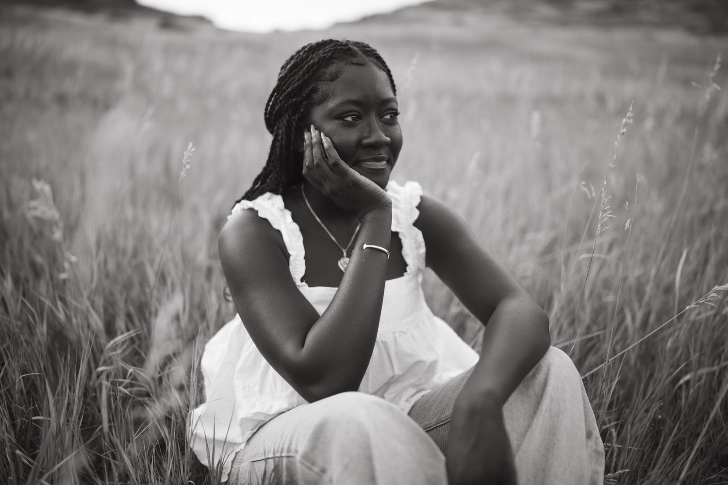 Black and white portrait of someone sitting thoughtfully in a field of tall grass wearing a white dress.