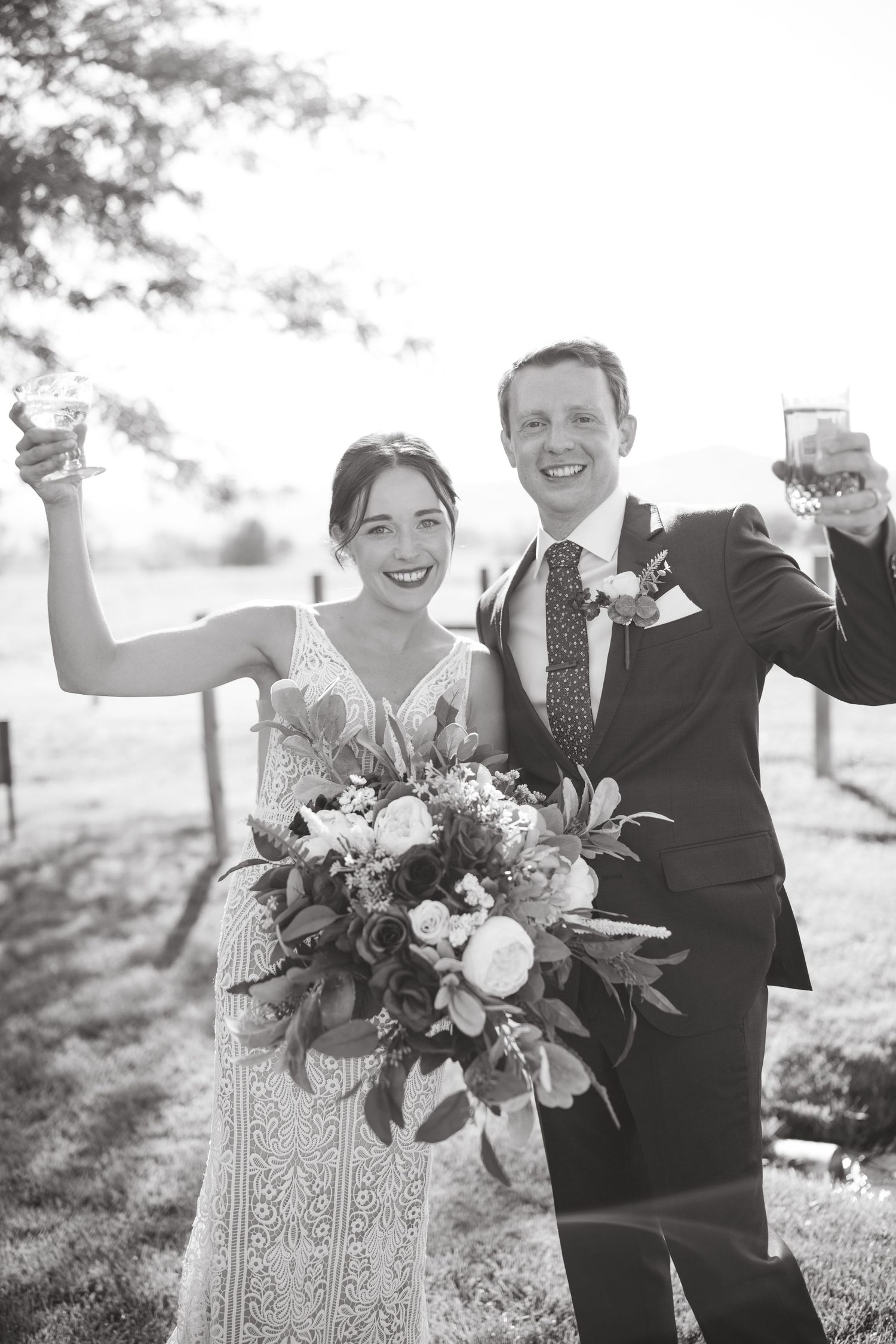 Joyful wedding couple celebrates with bouquet and raised arms in outdoor setting.