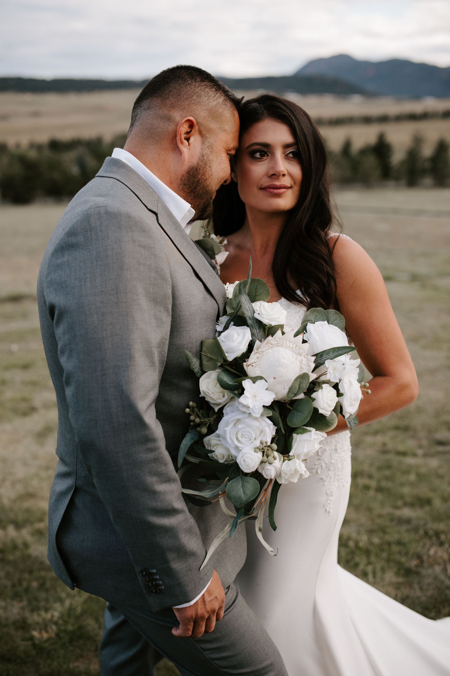 Elegant bride holding white rose bouquet stands with groom in grey suit against mountain landscape.