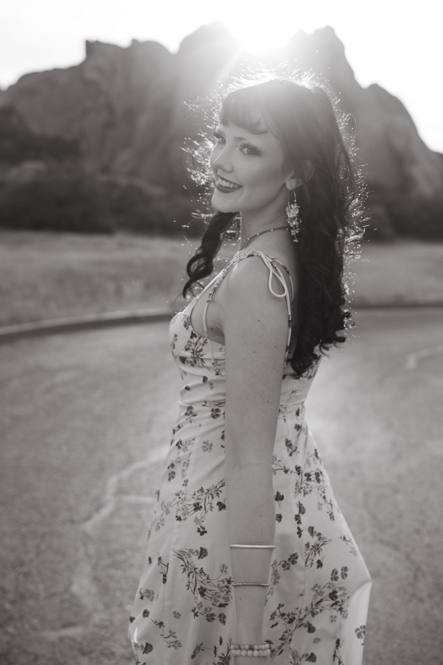 A woman in a floral dress laughs joyfully while standing in a sunlit field during golden hour.
