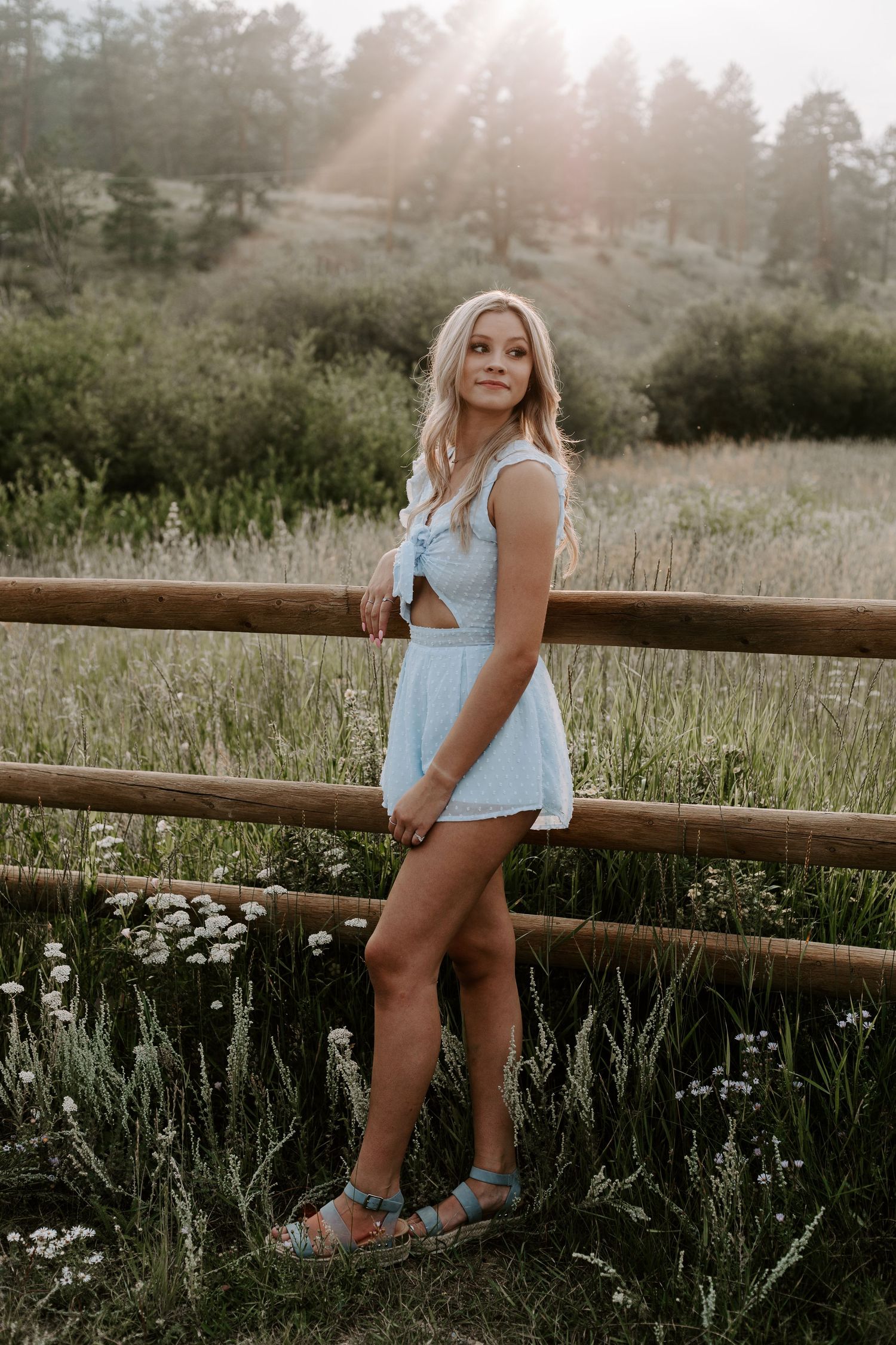 Golden hour portrait by rustic wooden fence wearing blue summer dress with mountains in background.