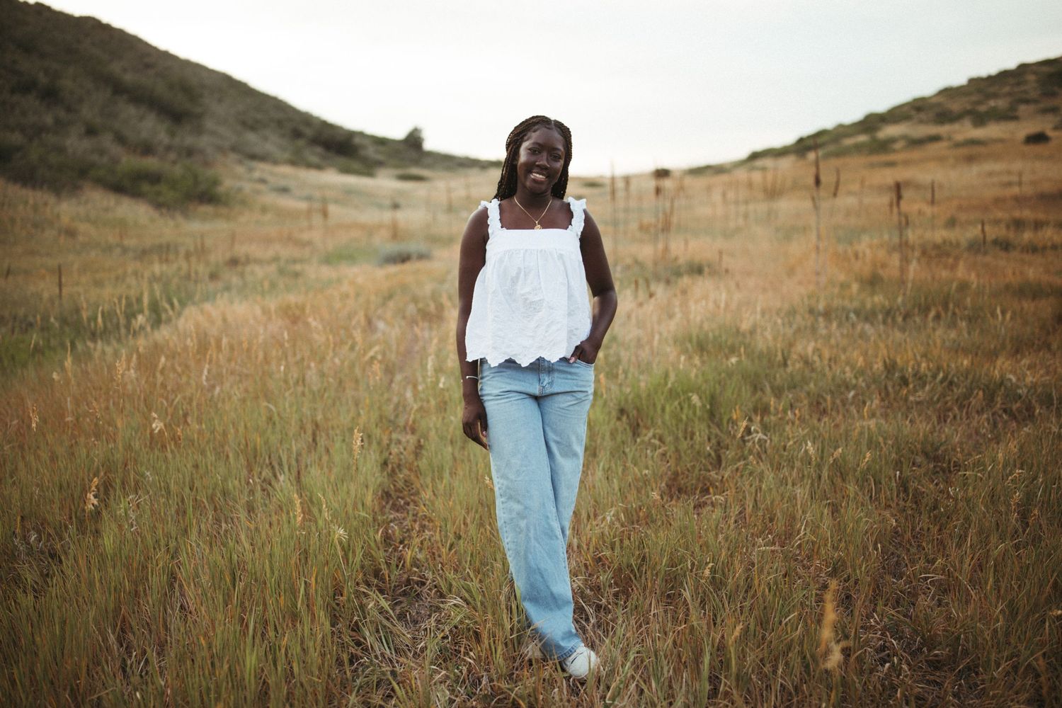 A person walks through golden hillside grass at sunset wearing light blue jeans and a white top.