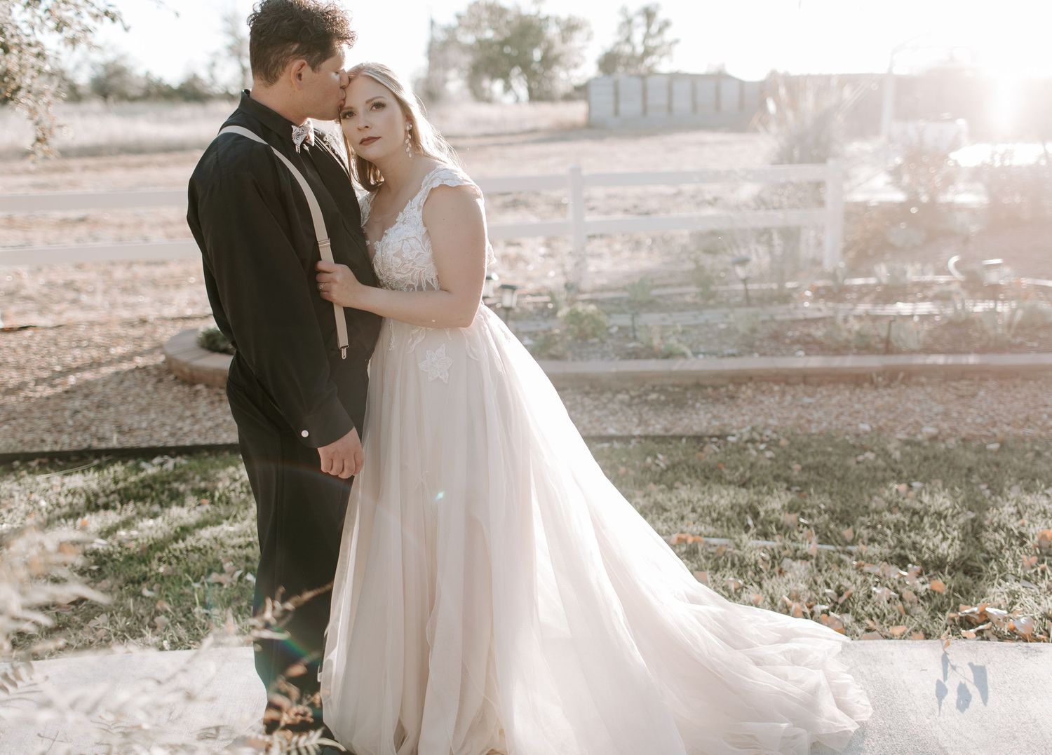 A newlywed couple embraces in a sunlit outdoor setting wearing formal wedding attire.