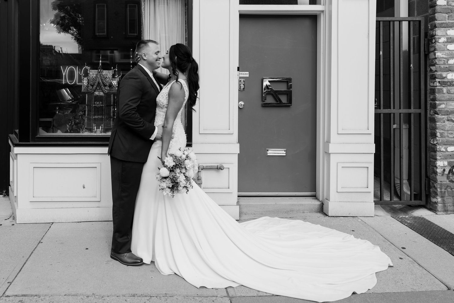Black and white wedding photo of a couple sharing a tender moment in front of a storefront.