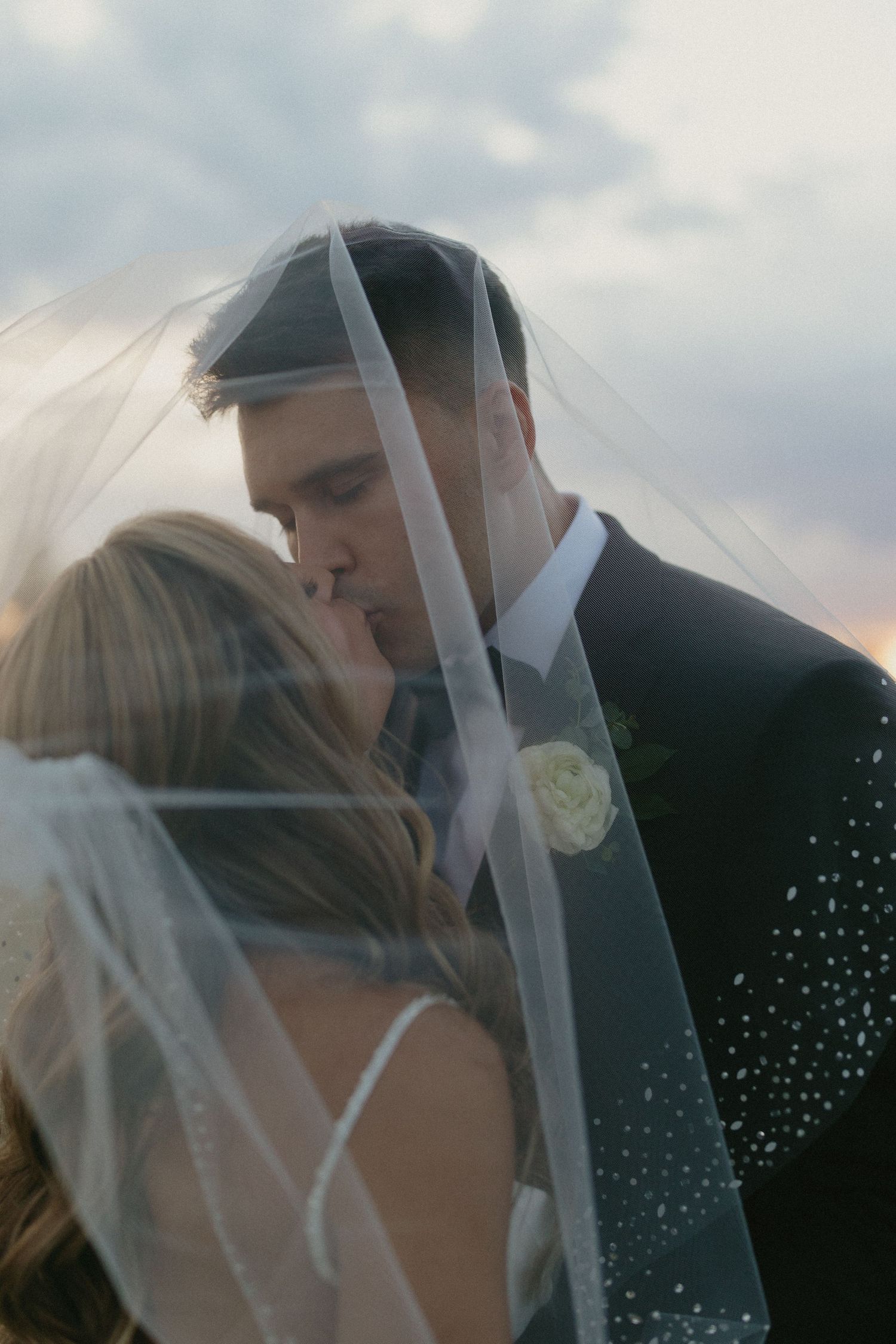 Bride and groom sharing a romantic kiss under a sheer veil at sunset on their wedding day.