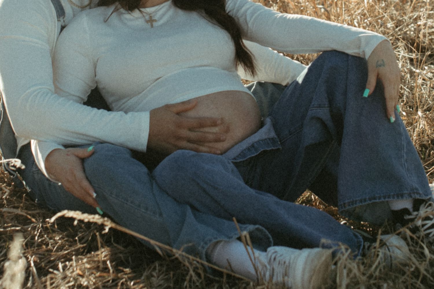 Close-up shot of hands resting on pregnant belly while couple sits in grassy field.