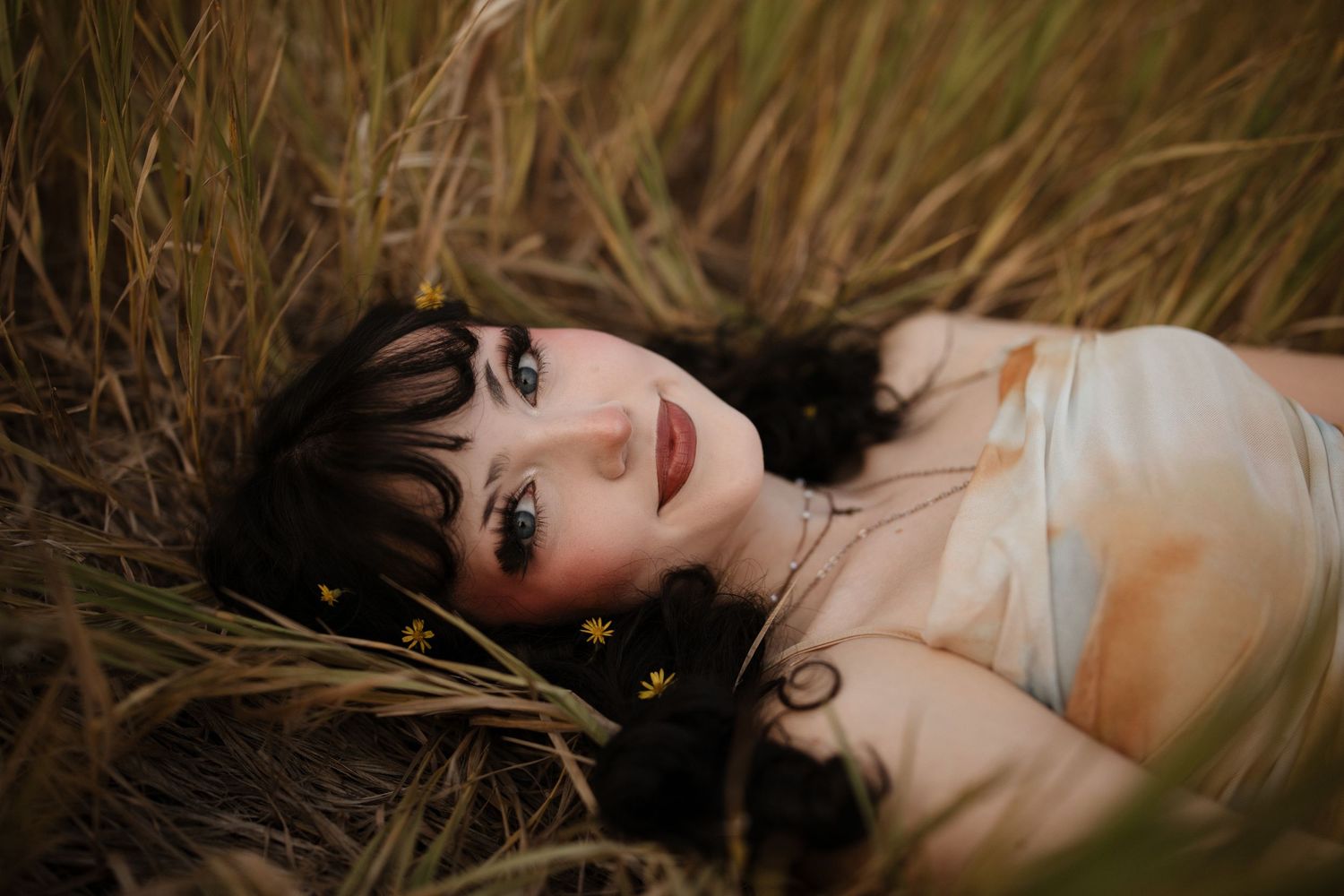 A person with dark hair lying in tall grass with a dreamy expression, creating a romantic outdoor portrait.