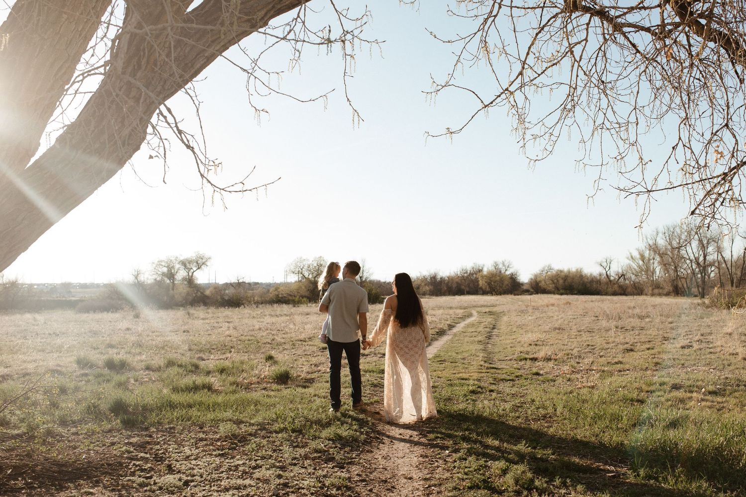 A couple walks hand in hand through a sunlit field under bare tree branches.