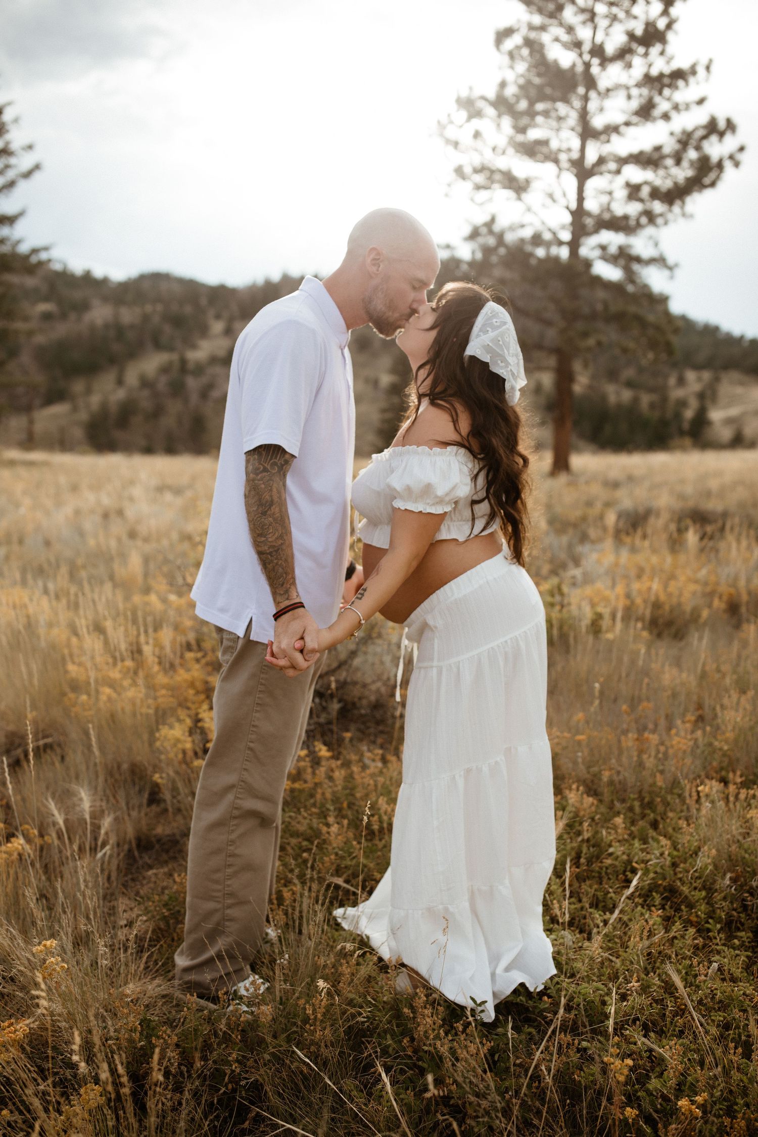 A romantic couple embraces in a golden field at sunset with tall pine trees in the background.