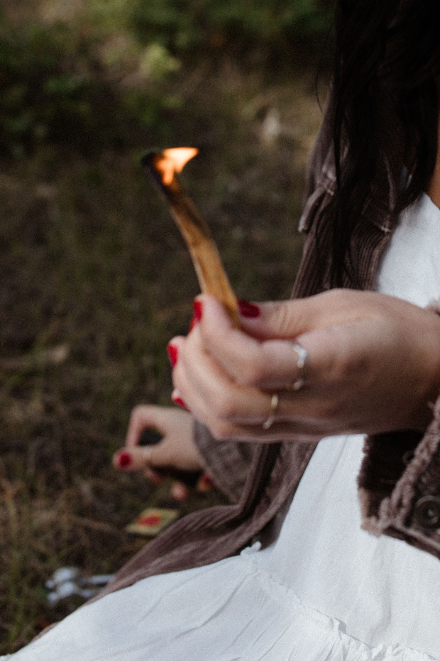 A person in white holding a lit match with red painted nails against a dark outdoor background.