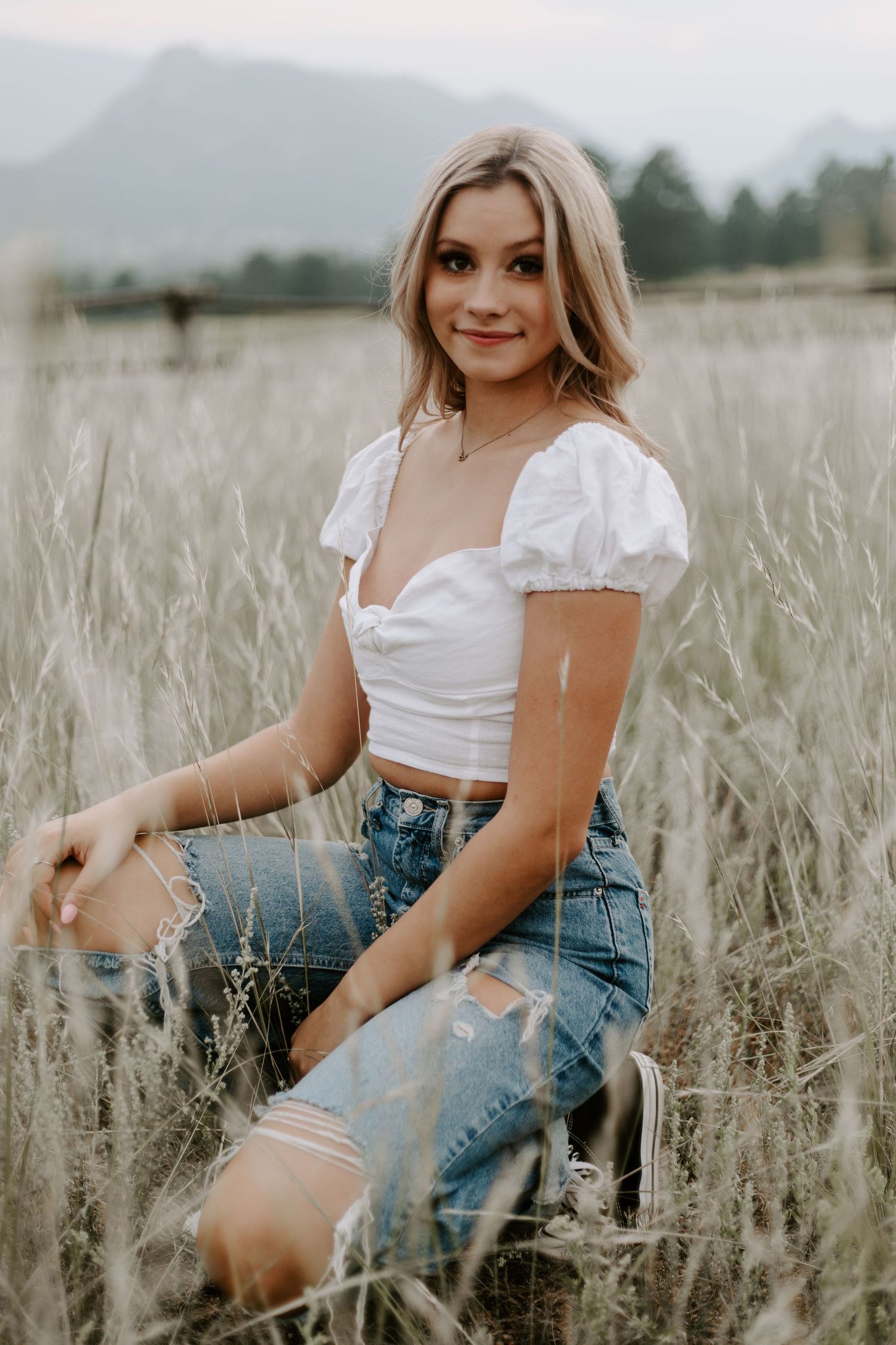 Summer portrait in a tall grass field with mountains in background wearing white top and ripped jeans.
