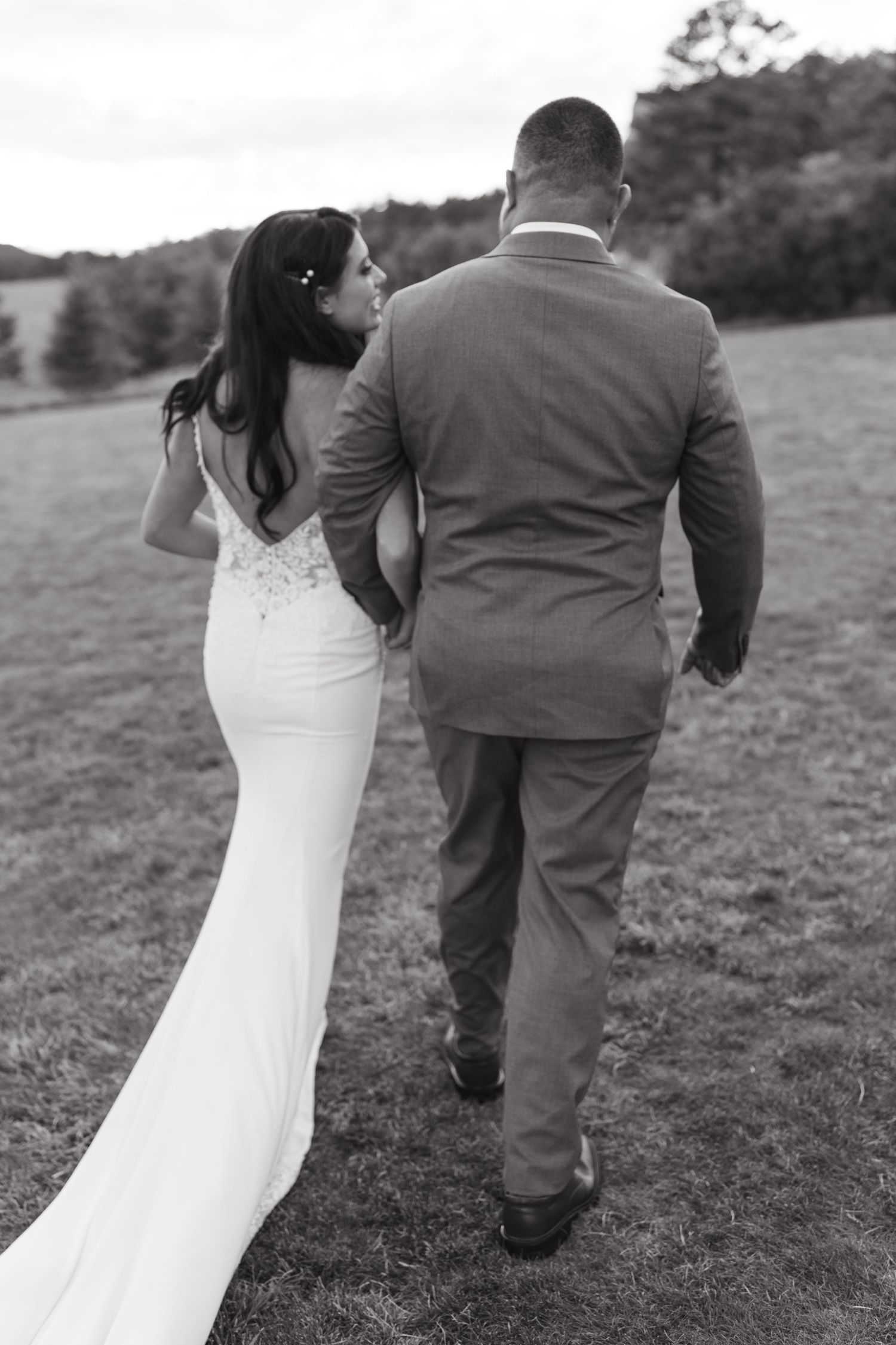 Black and white photo of newlyweds walking away together across mountainside meadow.
