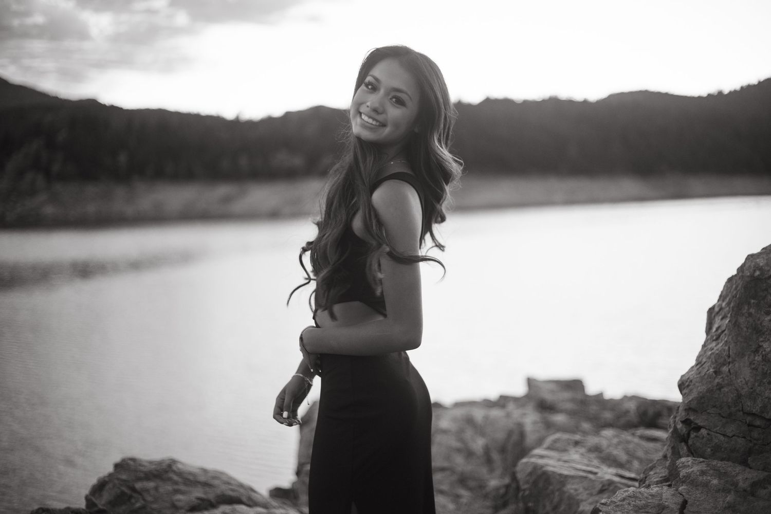 Black and white photo of woman in dark dress posing on rocks by lake.