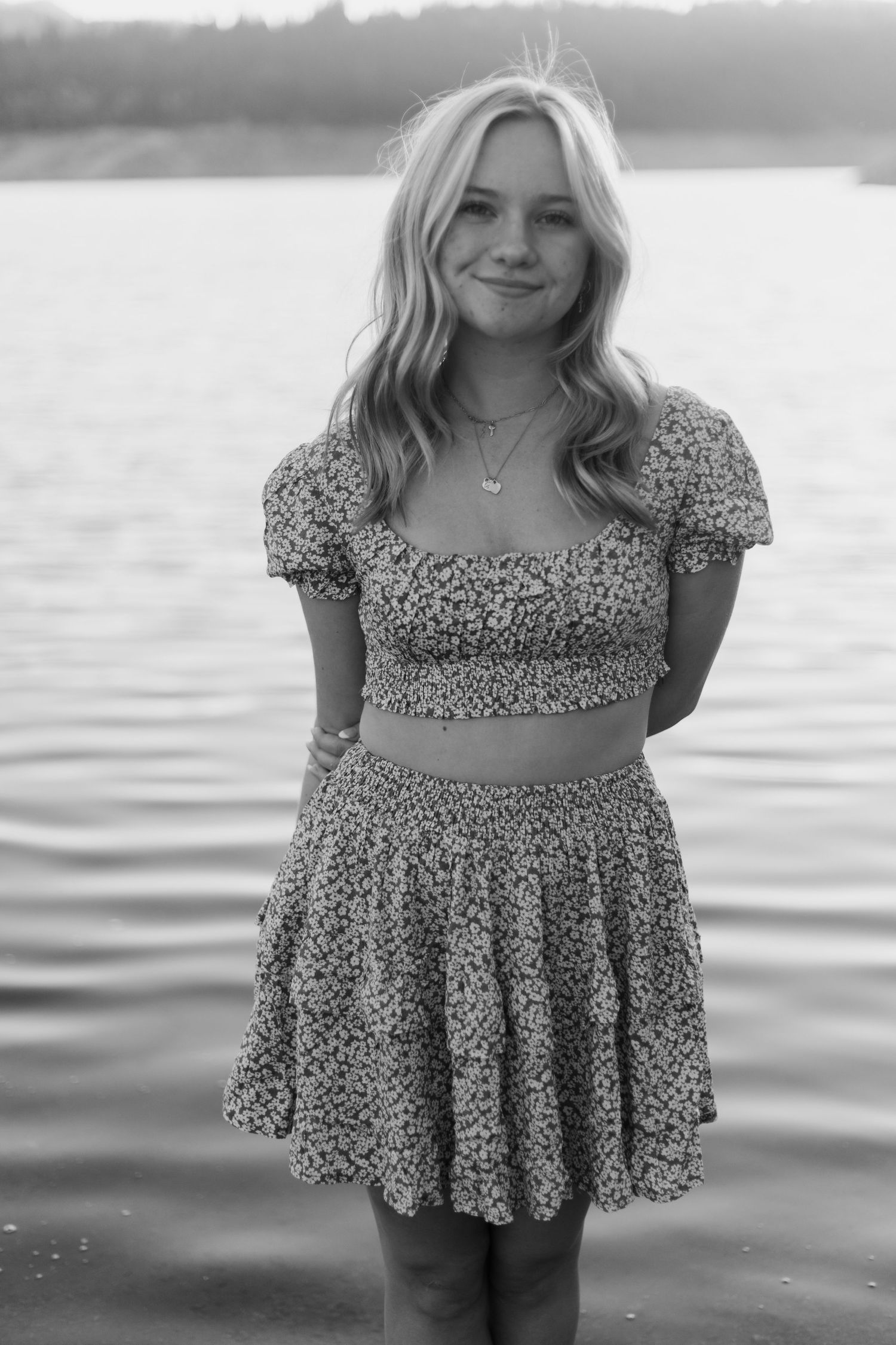 Person wearing floral summer dress poses near calm waterfront in black and white.