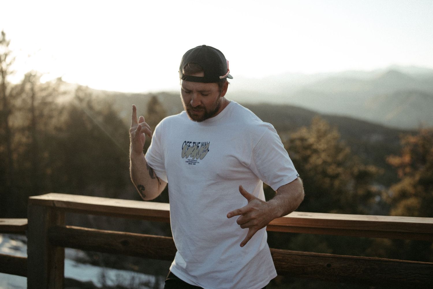 Person in white t-shirt stands at scenic mountain overlook during sunset while gesturing with hand.
