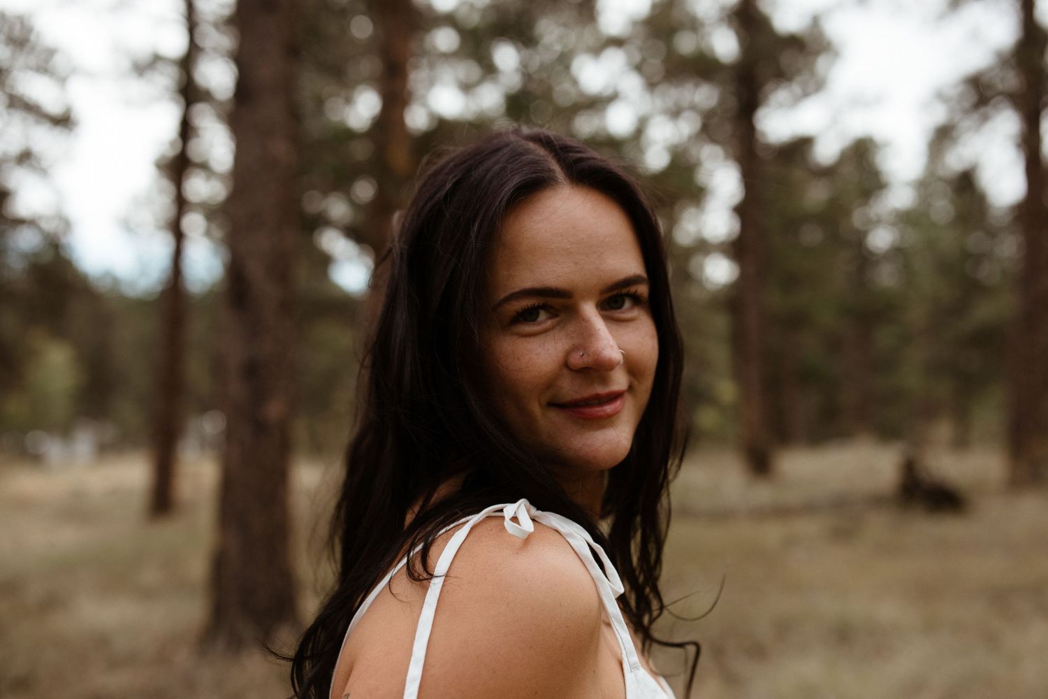 A person in a white dress poses outdoors in a wooded area with pine trees in the background.