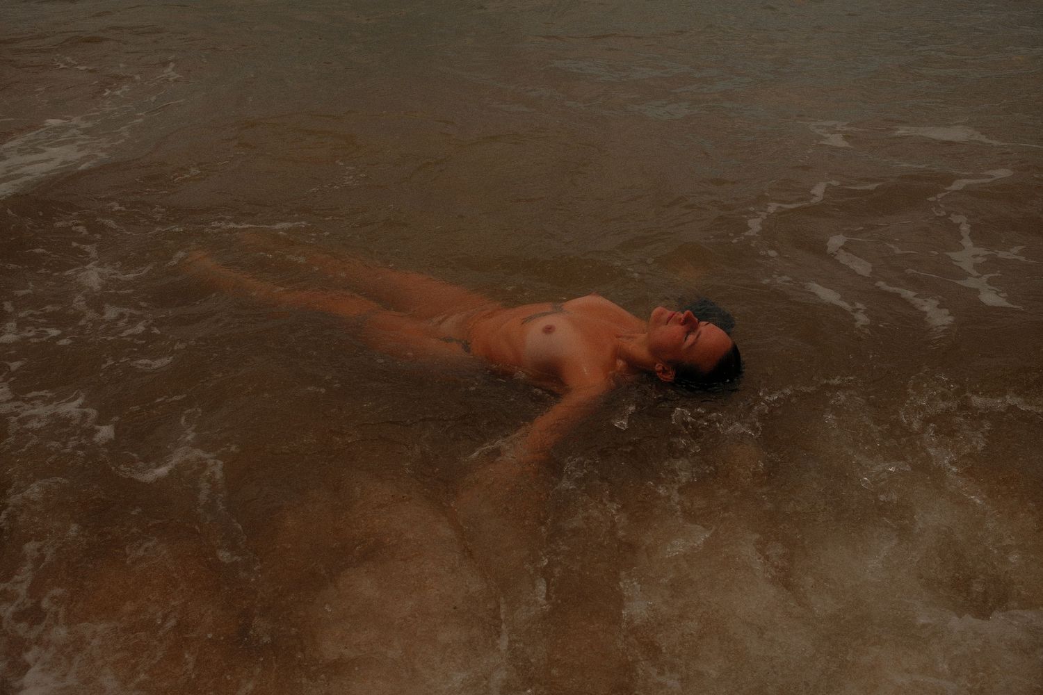 Person floating peacefully on their back in shallow ocean waves on a sandy beach.