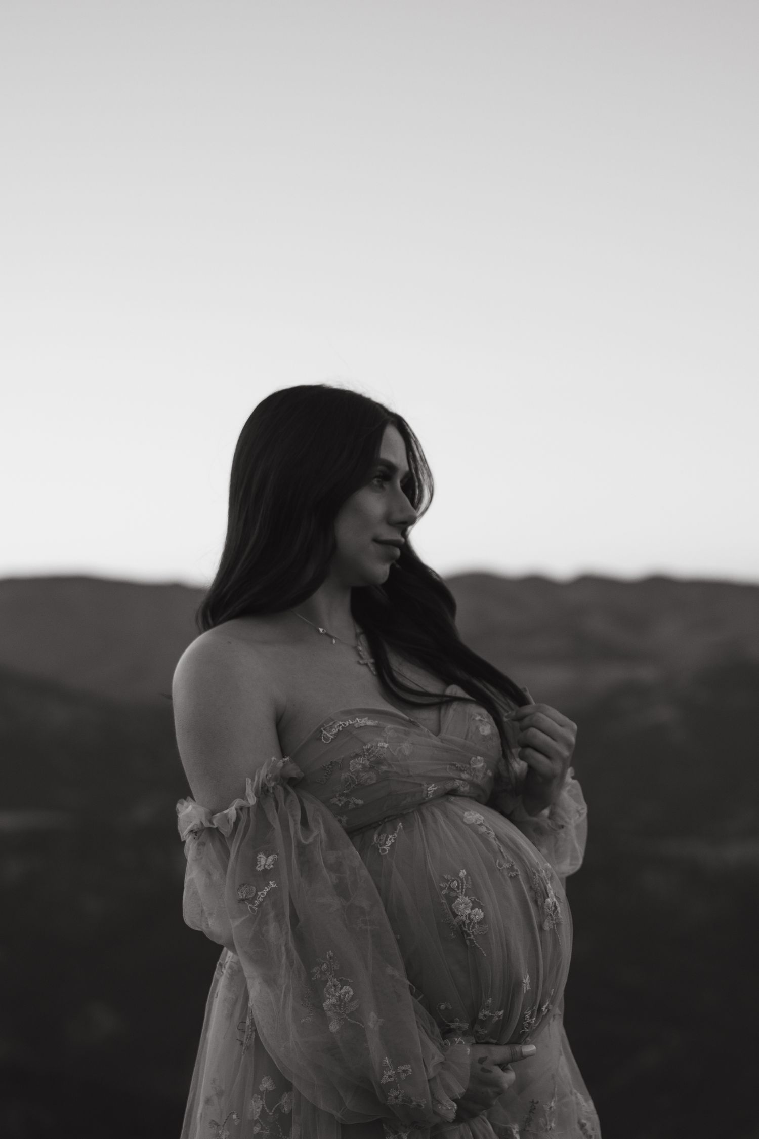 Black and white portrait of pregnant woman in floral dress against mountain landscape.