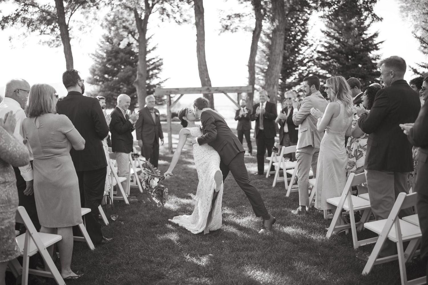 Wedding guests seated in white chairs at an outdoor ceremony venue surrounded by trees.