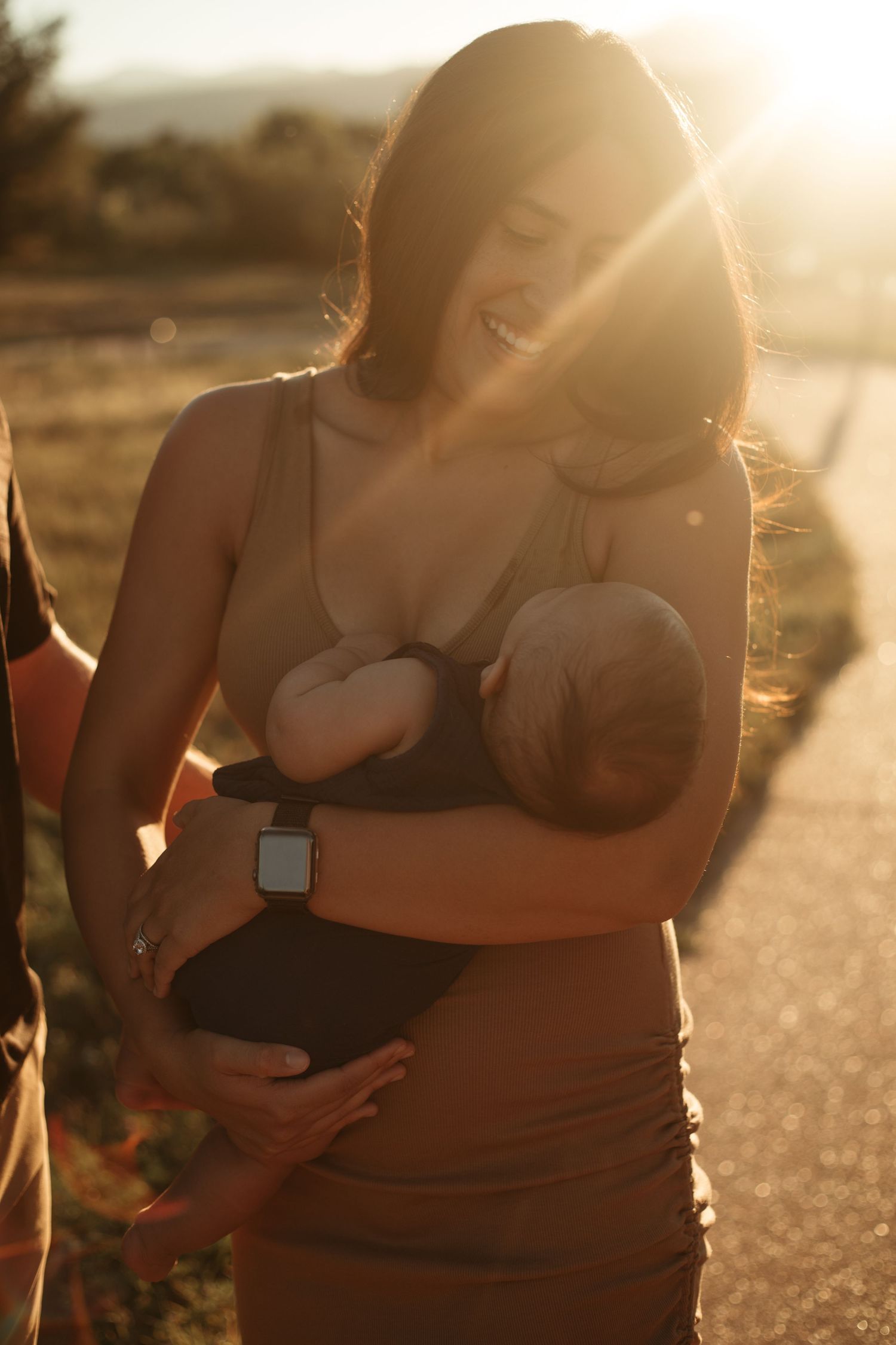 A parent cradling a newborn baby outdoors during golden hour sunset, backlit by warm sunlight.