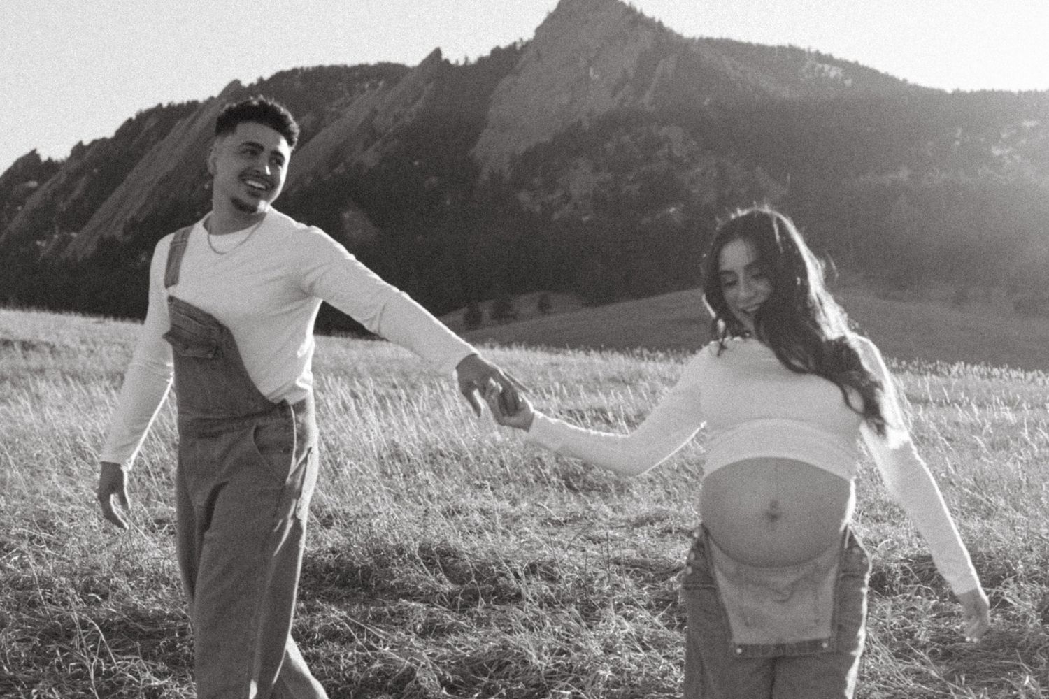 Black and white photo of expecting couple playfully holding hands in field with mountain backdrop.