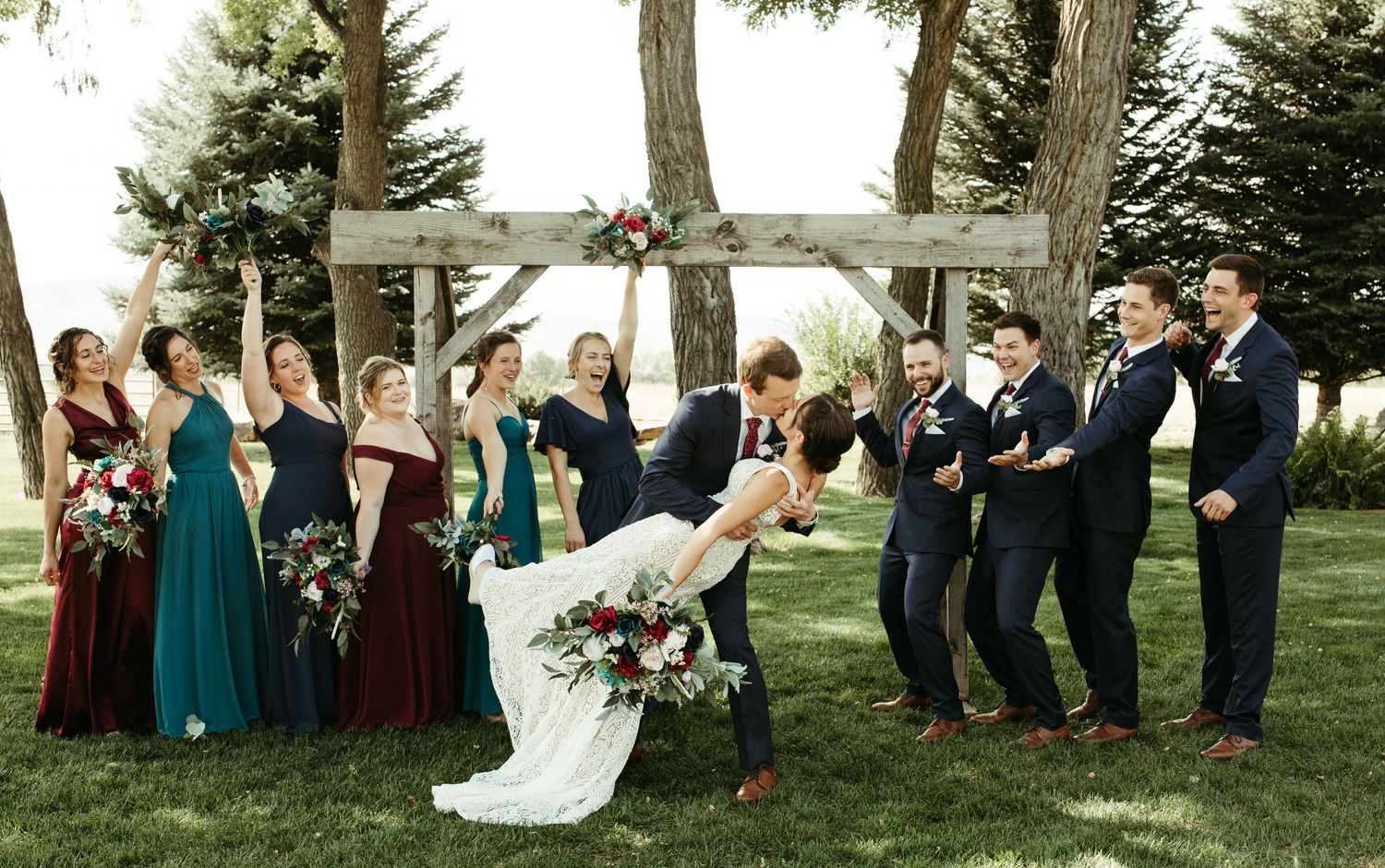 Wedding party poses for photos under large trees with bridesmaids in jewel-toned dresses.