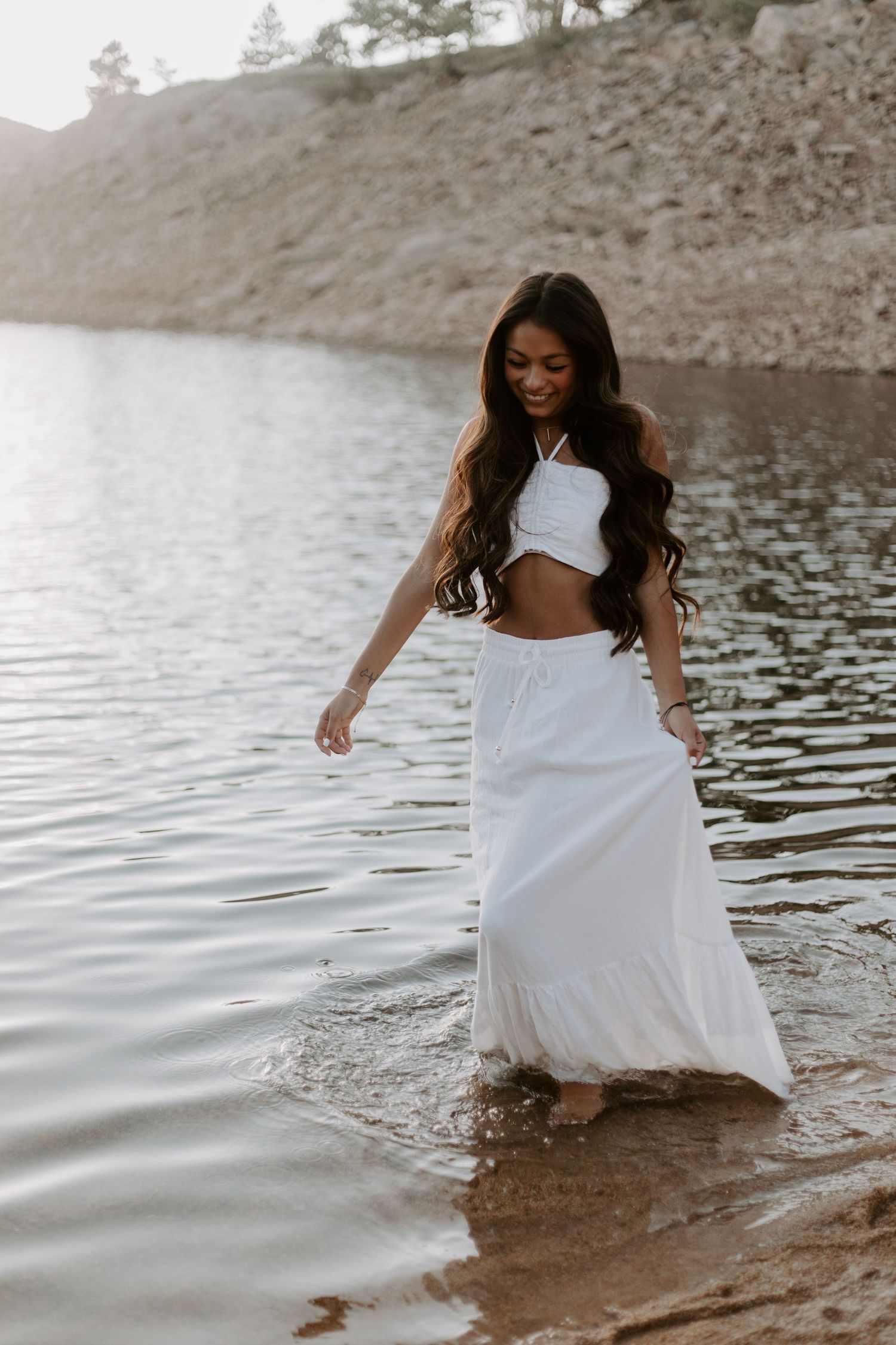Portrait at lake shore wearing white two piece outfit with rocky cliff in background.