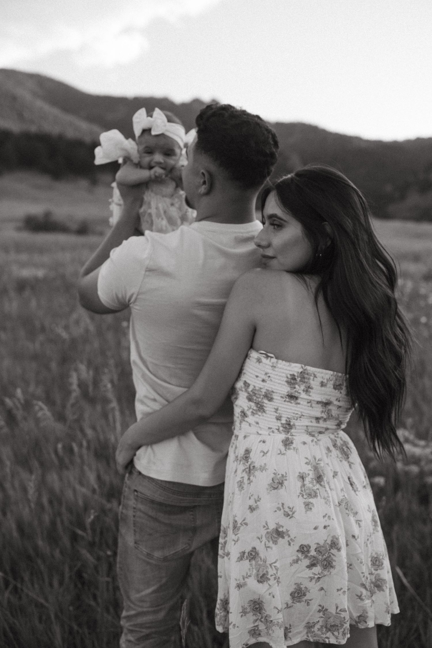 A romantic couple embraces in a field at sunset, with the woman wearing a floral sundress and man in casual attire.