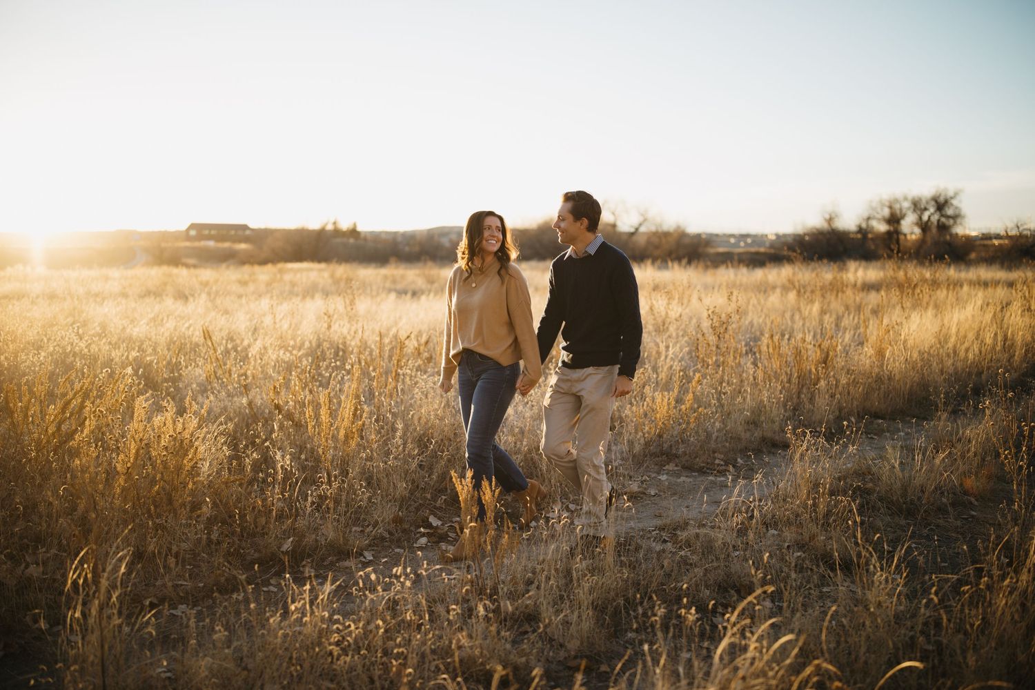 Couple walking together through a golden field at sunset with mountains in background.