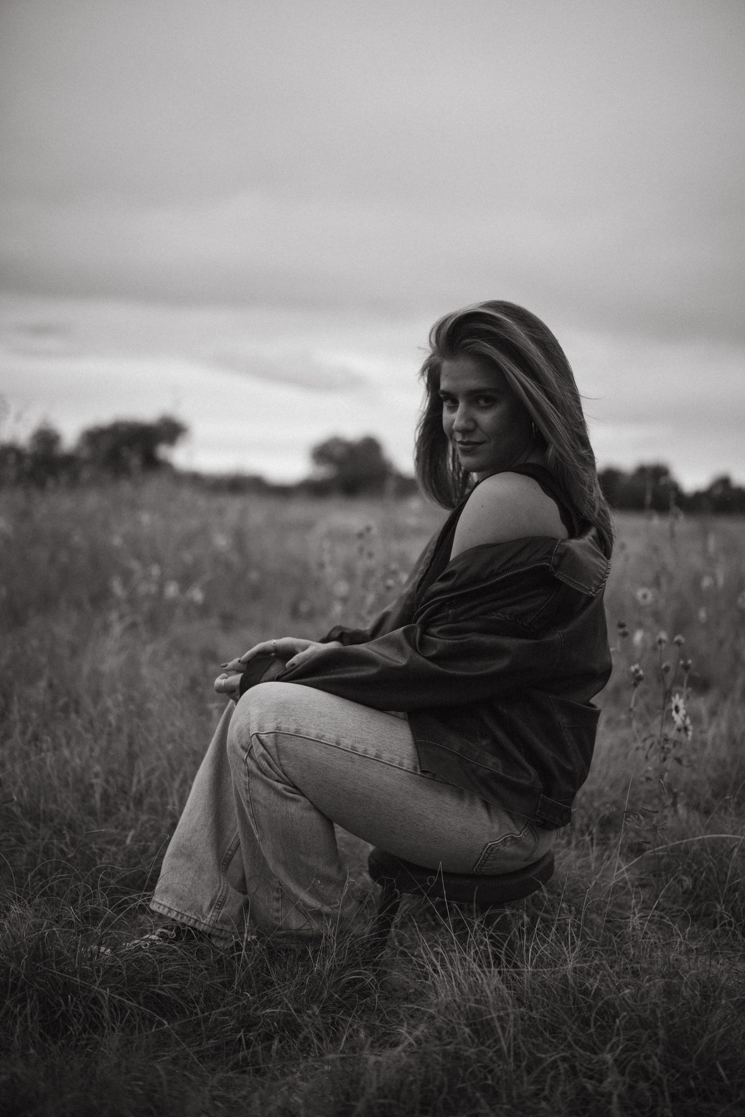 A figure in dark clothing sits thoughtfully in a grassy meadow on an overcast day.