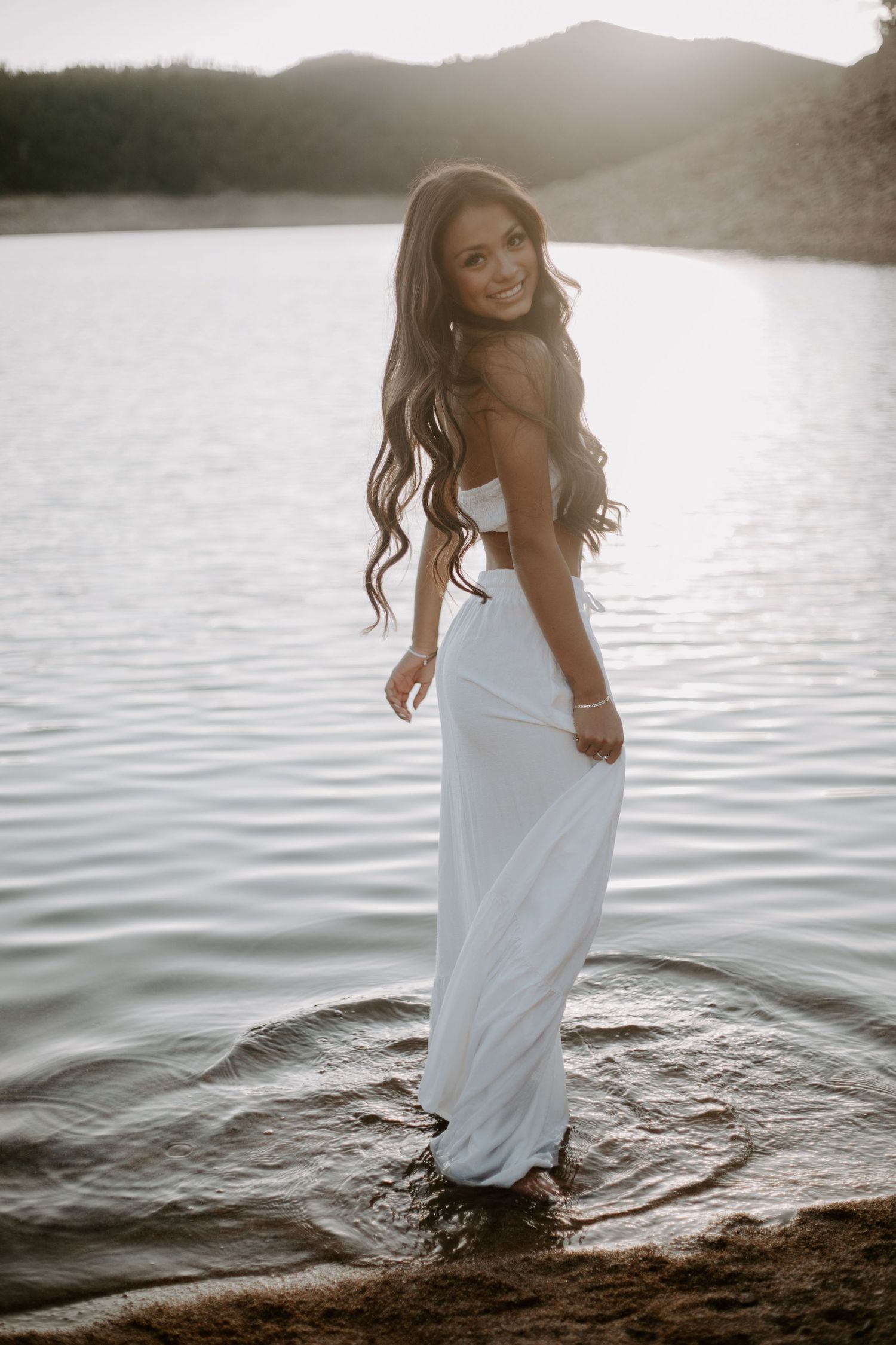 Woman in white dress poses gracefully by a serene lake with mountains in background.